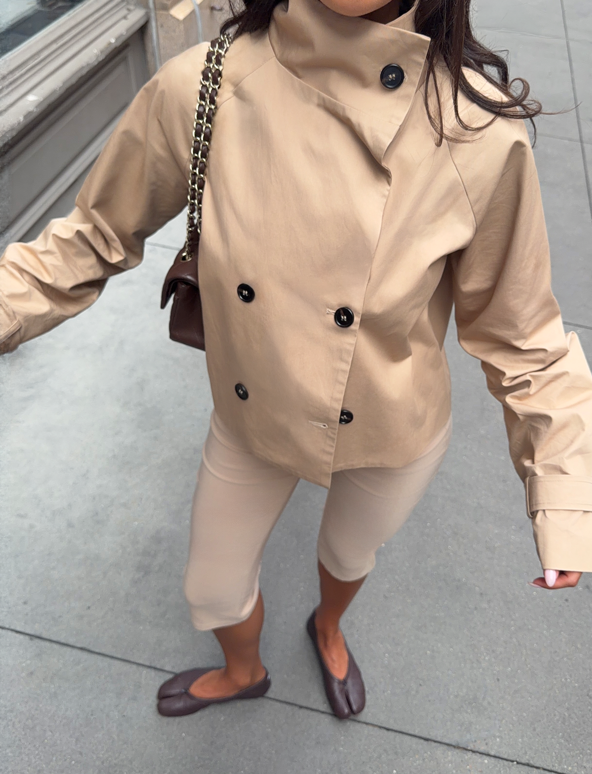 A person wearing the TIGERMIST HAYLEY CROPPED TRENCH JACKET in khaki, paired with beige pants and brown flats, stands on a city sidewalk carrying a chain-strap shoulder bag. The photo is taken from above, capturing only the lower part of their face.