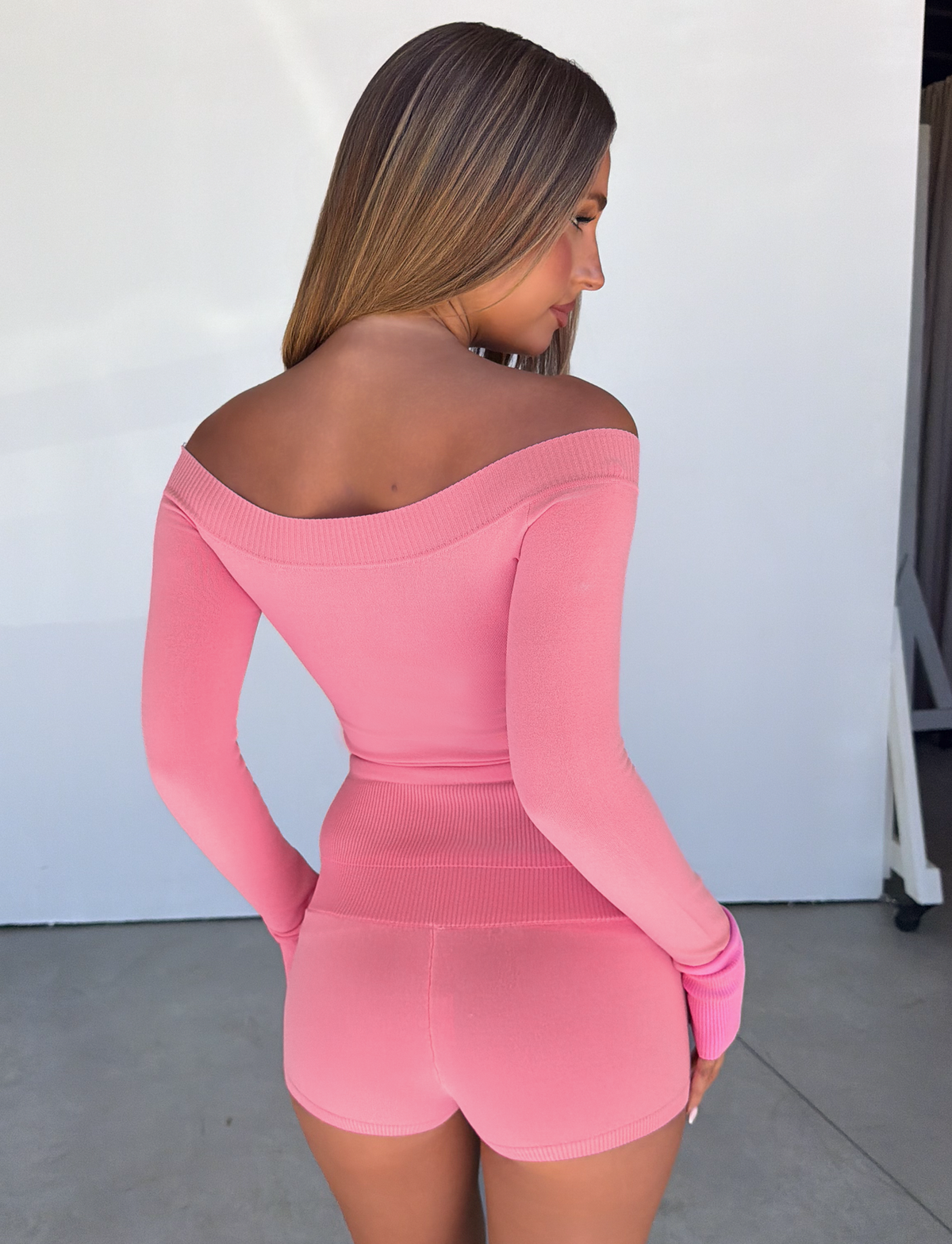 A woman with long hair stands with her back to the camera in a brightly lit space, wearing a pink off-the-shoulder top and TIGERMIST PARKER KNIT MICRO SHORTS in coral, featuring a bodycon fit.