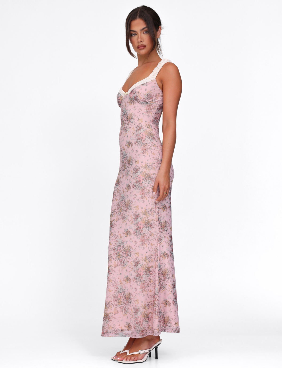 A woman in profile wears the TIGERMIST AMARAN DRESS - PINK : CAMO FLORAL, featuring thin straps and a maxi silhouette. Her dark hair is styled up, paired with white heeled sandals against a plain white background.