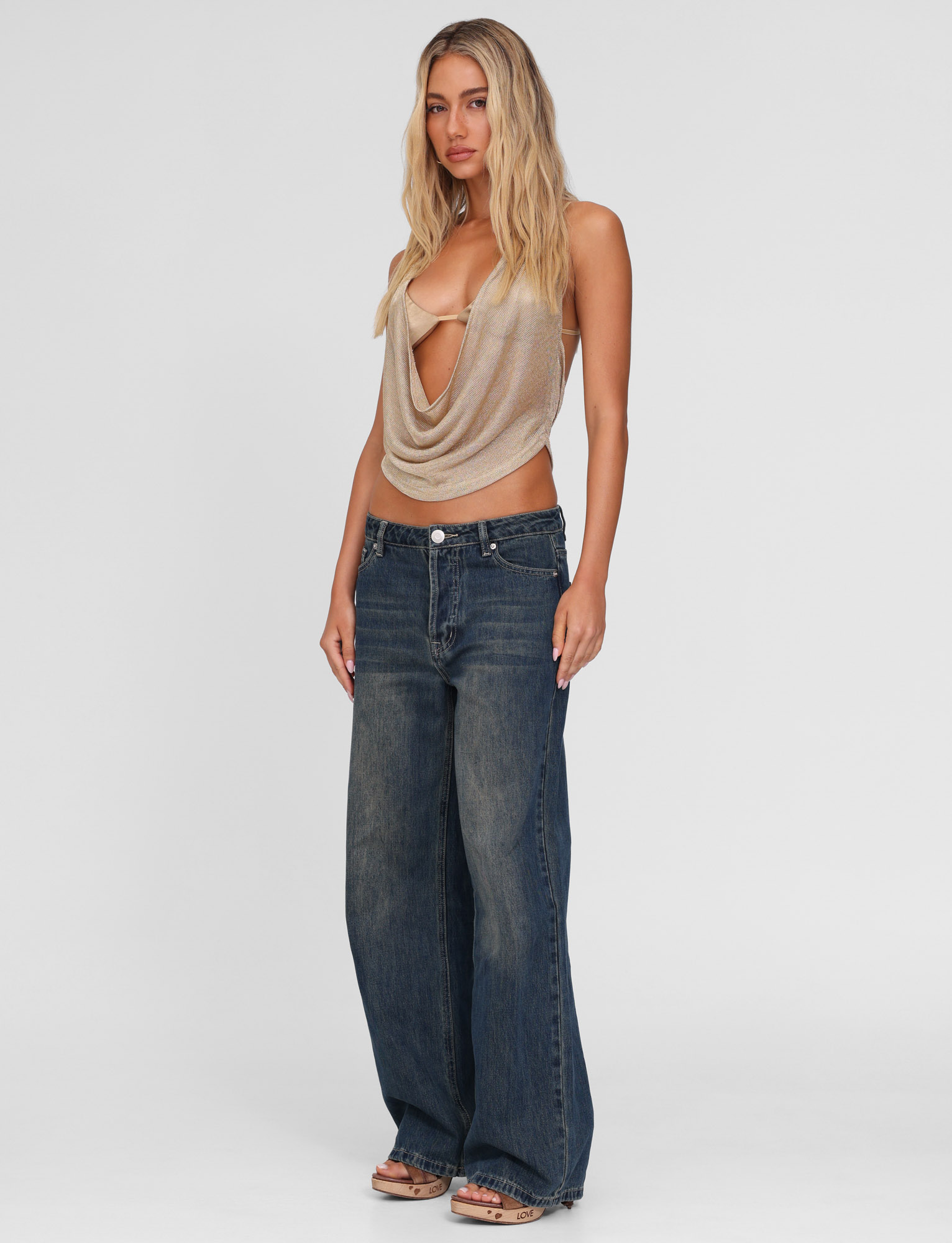 A woman with long, wavy blonde hair wears the TIGERMIST FELIX JEAN - BLUE : MID WASH and a beige halter top, standing front-facing against a plain white background with a neutral expression.