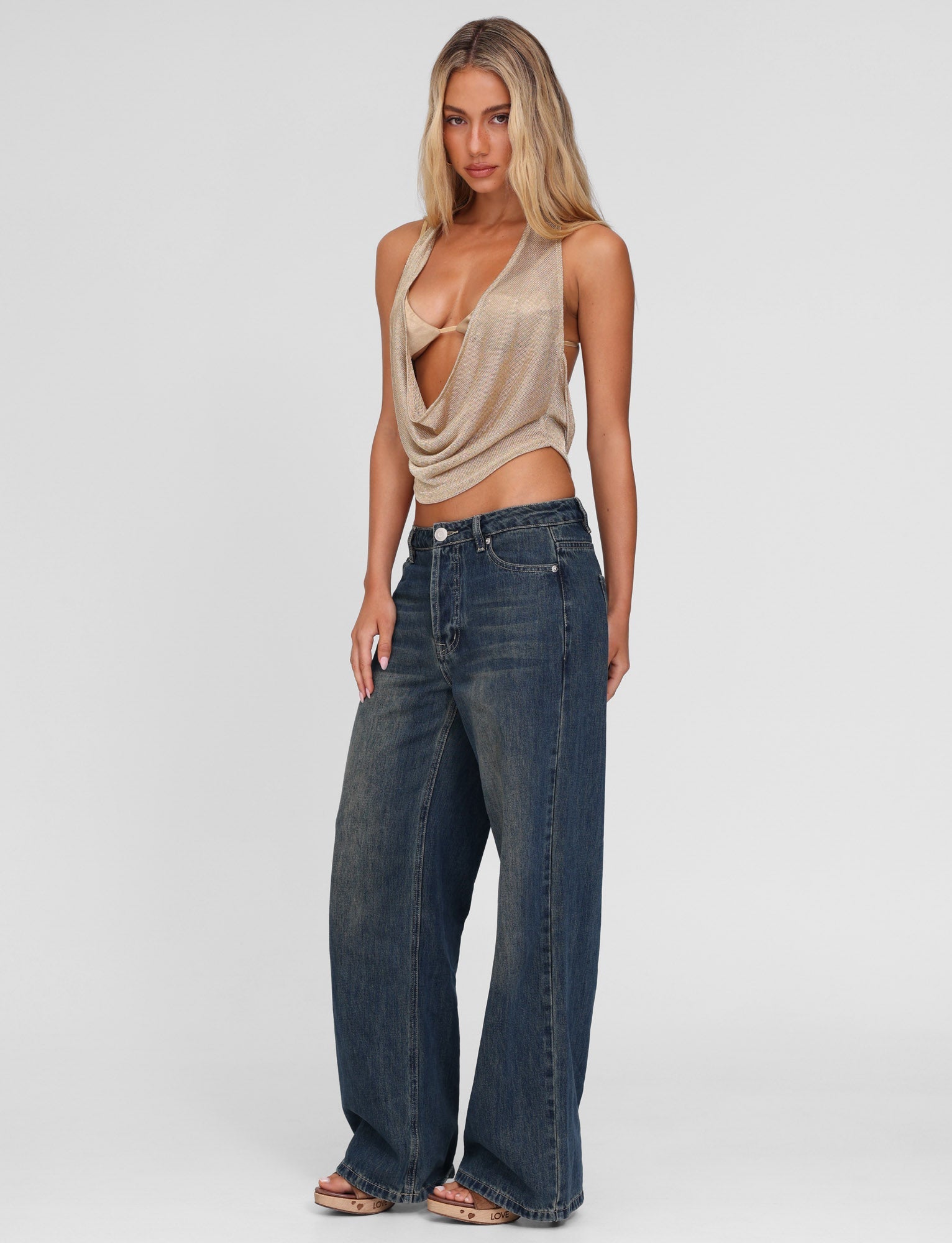 A woman with long, wavy blonde hair stands against a plain background wearing TIGERMIST's FELIX JEAN - BLUE: MID WASH, styled high-waisted and wide-leg, paired with a beige draped halter top. She looks directly at the camera with a neutral expression.