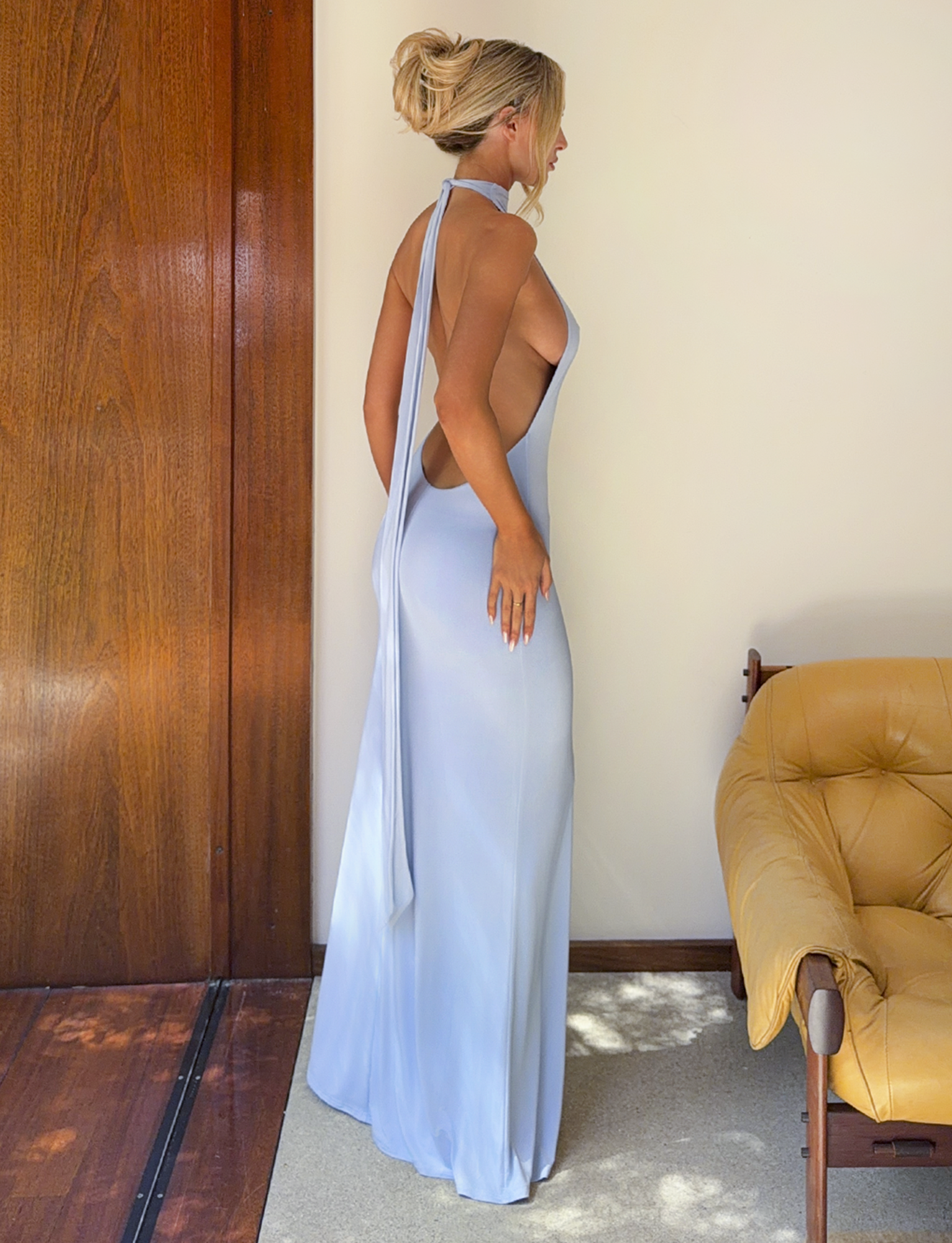 A woman with blonde hair in an updo wears the TIGERMIST MADDY MAXI DRESS in baby blue, standing sideways by a tan armchair and wooden cream-colored walls, looking away from the camera.