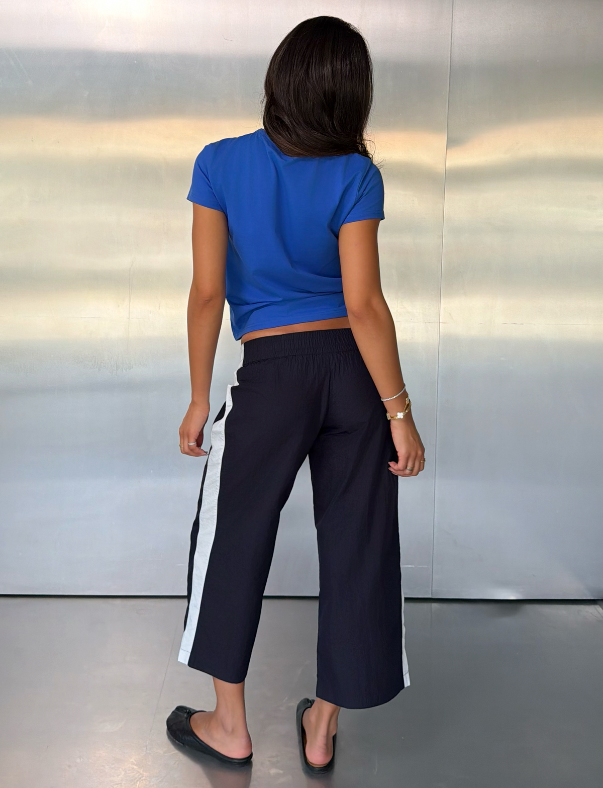 A woman stands indoors facing away, wearing a blue t-shirt, black flats, and the TIGERMIST SOPHIA WINDBREAKER CAPRI in navy and white with wide side stripes. The background is a smooth metallic wall.