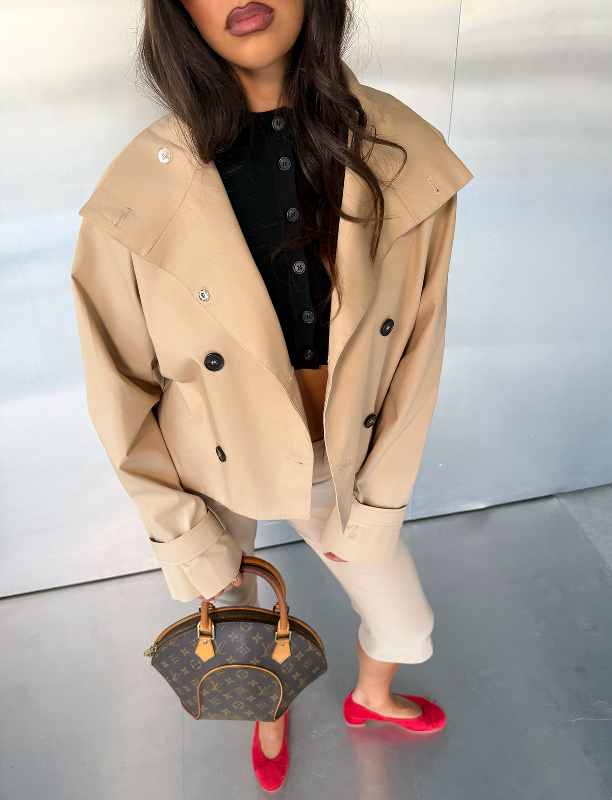 A woman wearing the HAYLEY CROPPED TRENCH JACKET in khaki by TIGERMIST, paired with a black top, beige pants, and red shoes, holds a brown designer handbag while standing on a gray floor against a light-colored background.