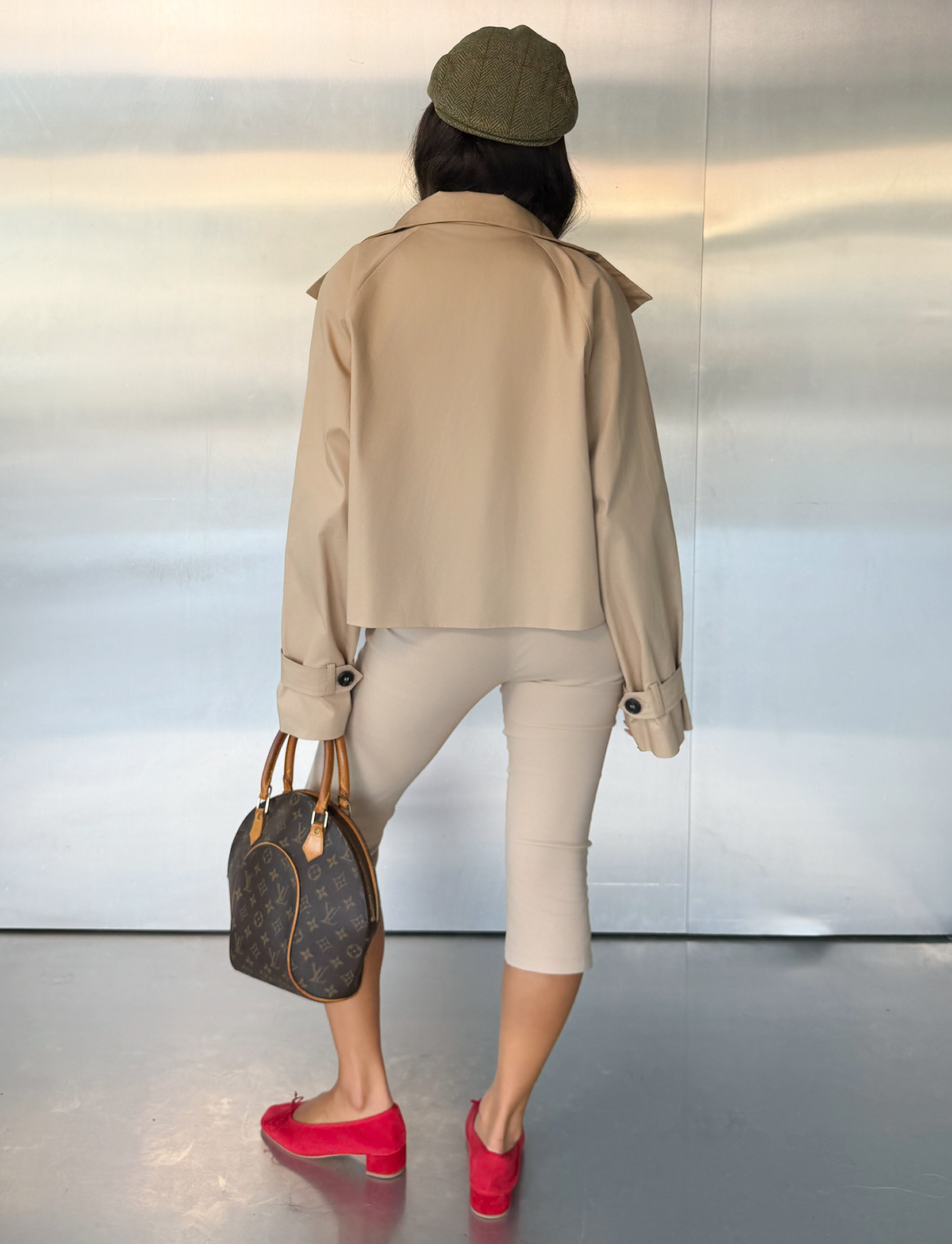 A person stands facing away, wearing the HAYLEY CROPPED TRENCH JACKET in khaki from TIGERMIST, paired with matching capri pants, a green cap, red loafers, and holding a brown designer handbag. The background features a smooth, metallic wall.
