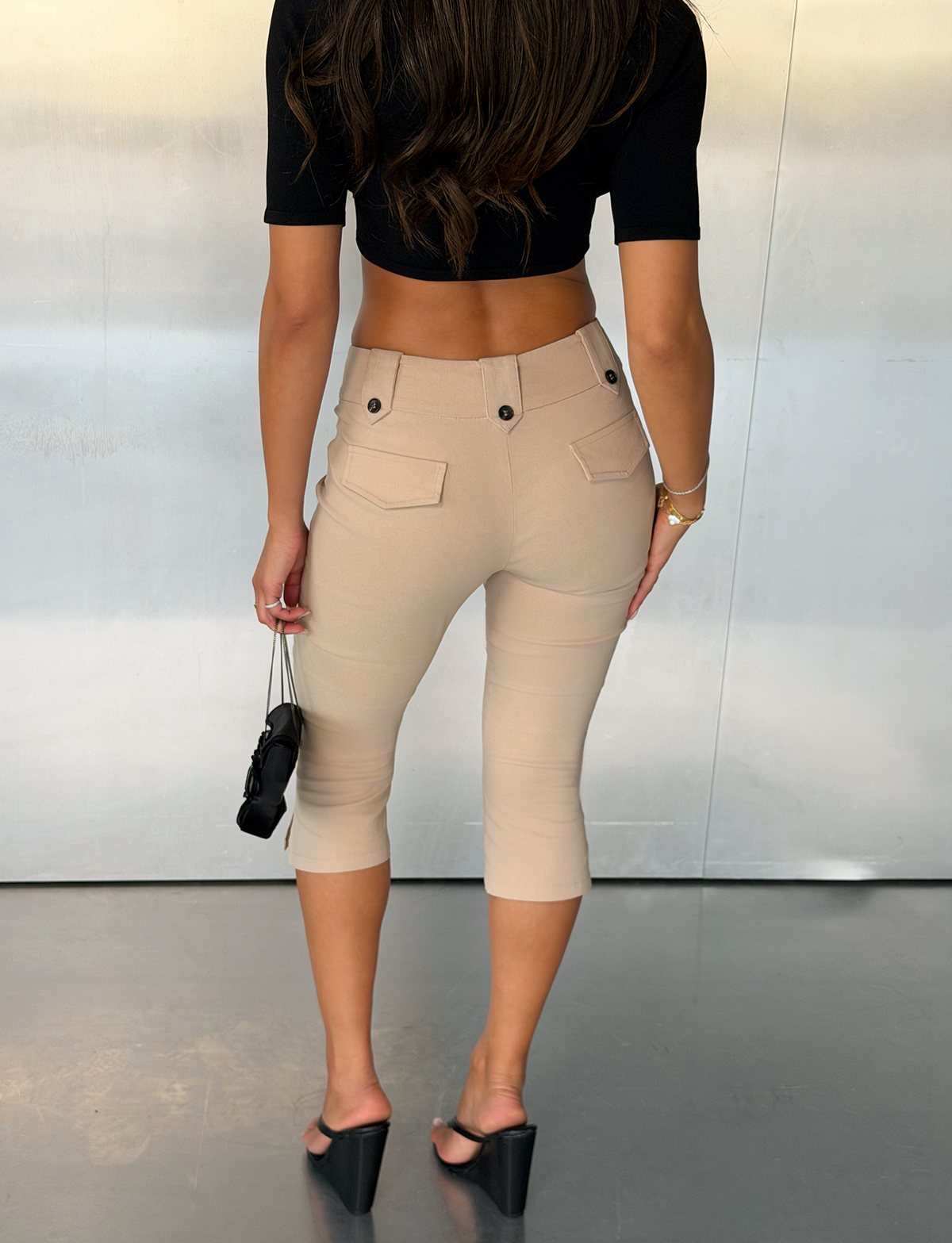 A woman stands indoors facing away, wearing a black crop top, the TIGERMIST ARIA CAPRI - KHAKI tailored fit mid-rise capri pants, and black wedge sandals, holding a small black purse in her left hand.