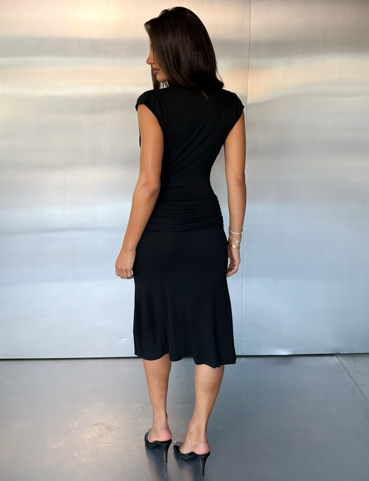 A woman with long brown hair wears the TIGERMIST ARIELLA PLUNGE MIDI DRESS in black and black heels, standing indoors on a polished floor, facing away from the camera against a light gray wall.