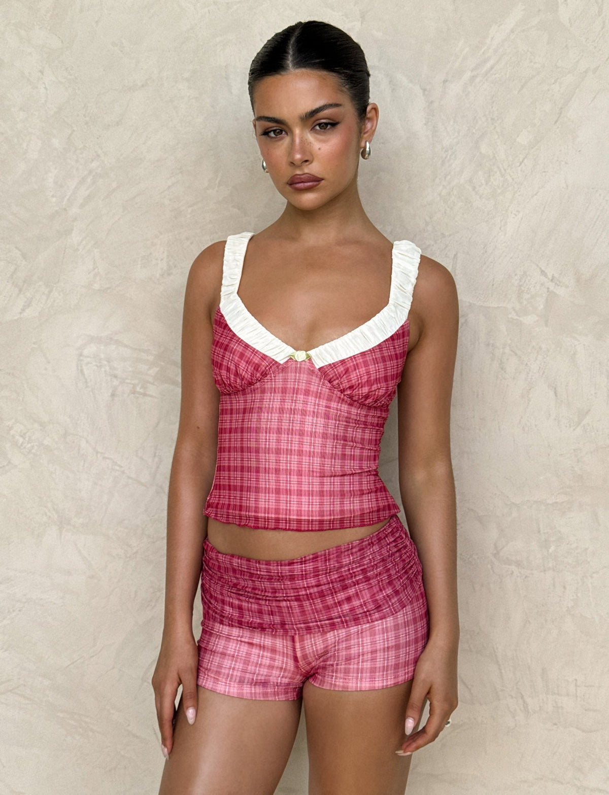 A person stands against a textured beige wall wearing a TIGERMIST AMARAN TOP - RED CHECK, featuring a white ruffled neck detail. They have slicked-back hair, hoop earrings, and a calm expression.