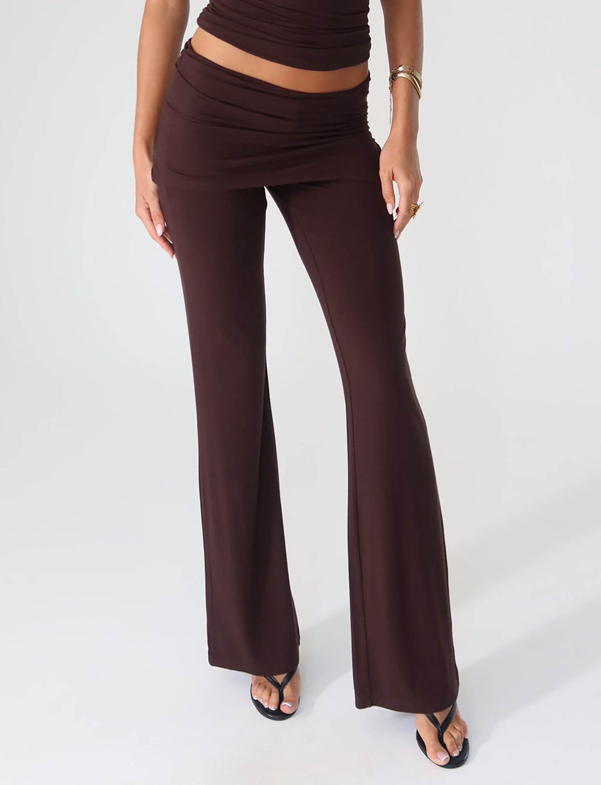 A person is standing against a light background, wearing the TIGERMIST LEILANI PANT in brown chocolate with a matching top. The high-stretch fabric of the pants features a layered waistband design. They are also wearing black sandals and a bracelet.