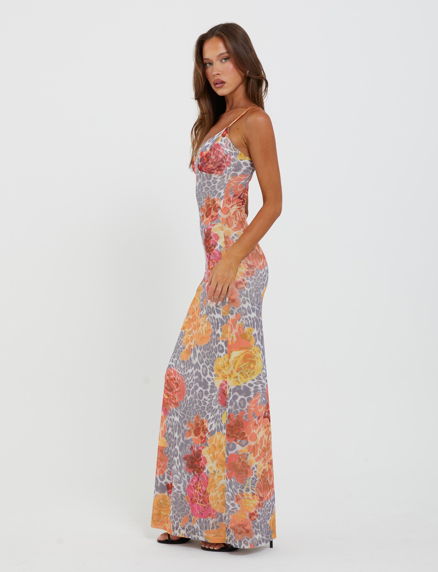 A woman is standing and modeling the Julianna Dress - Multi : Leopard : Orange Leopard by TigerMist. The sleeveless, body-hugging, floor-length dress showcases a vibrant floral and geometric pattern in shades of orange, yellow, pink, and gray. She has long brown hair and is posing sideways with one hand resting on her hip.
