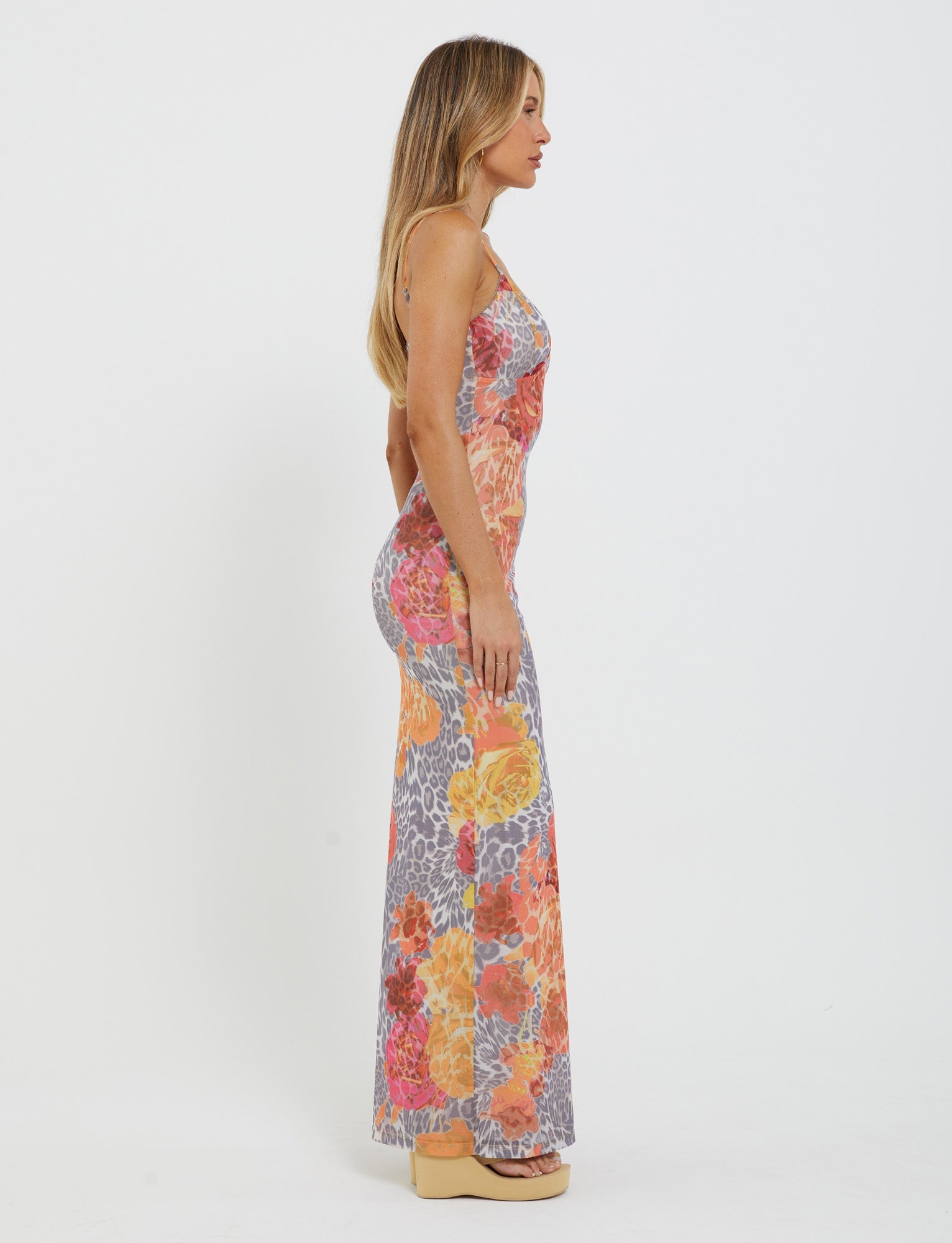 A woman with long blonde hair stands in profile against a plain white background. She is wearing the JULIANNA DRESS - MULTI : LEOPARD : ORANGE LEOPARD from TIGERMIST, a sleeveless, floor-length dress featuring a colorful floral and leopard print design. She complements her outfit with beige platform shoes.