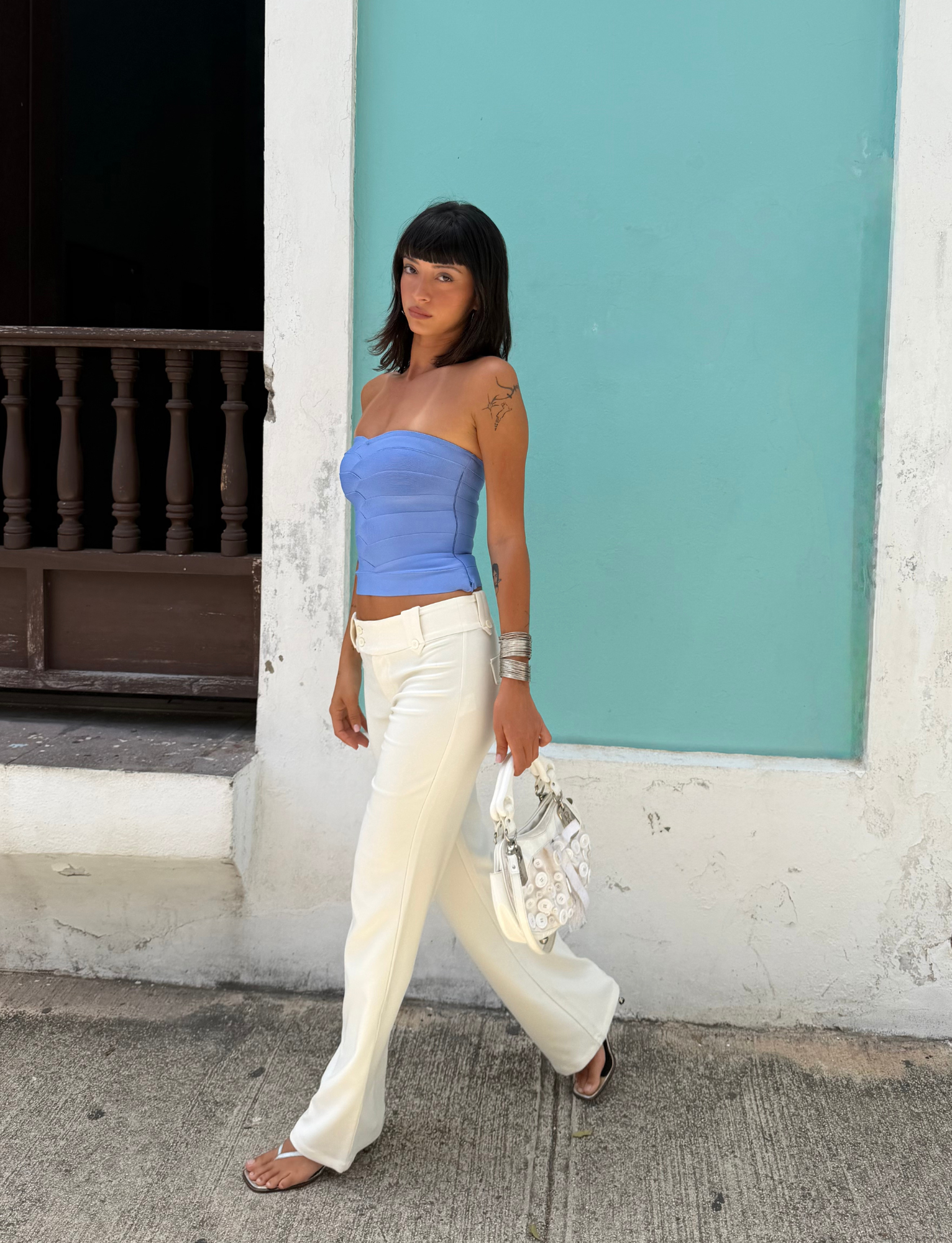 A woman with dark hair walks on a sidewalk in front of a turquoise wall and wooden railing, carrying a white purse. She wears the TIGERMIST NIYA BANDAGE TOP in cornflower blue, featuring a strapless, sweetheart neckline that pairs stylishly with her white pants.