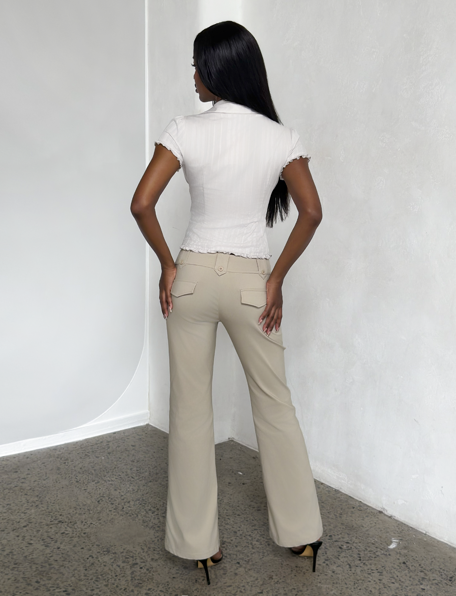 A woman with long dark hair stands facing a white textured wall, wearing a short-sleeve white blouse, TIGERMIST KITTIE PANT in sand stretch suiting fabric, and black high heels. The photo is taken from behind on a speckled floor.