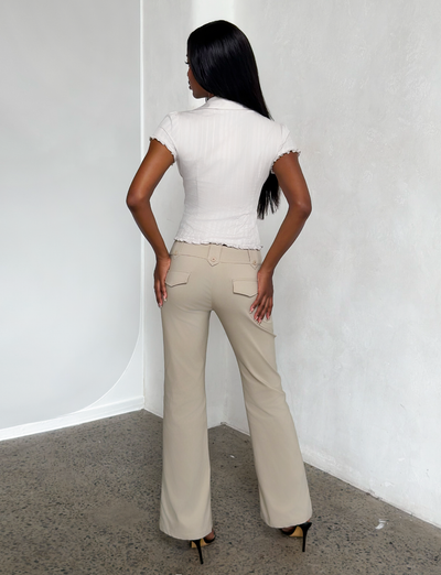 A woman with long dark hair stands facing a white textured wall, wearing a short-sleeve white blouse, TIGERMIST KITTIE PANT in sand stretch suiting fabric, and black high heels. The photo is taken from behind on a speckled floor.