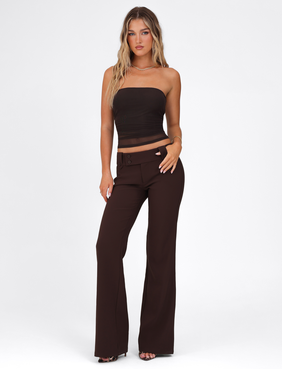 A woman with long wavy hair poses against a white background, wearing the TIGERMIST HARRIETT TUBE TOP - BROWN: CHOCOLATE with matching high-waisted, wide-leg pants. She accessorizes with a necklace and heeled sandals.