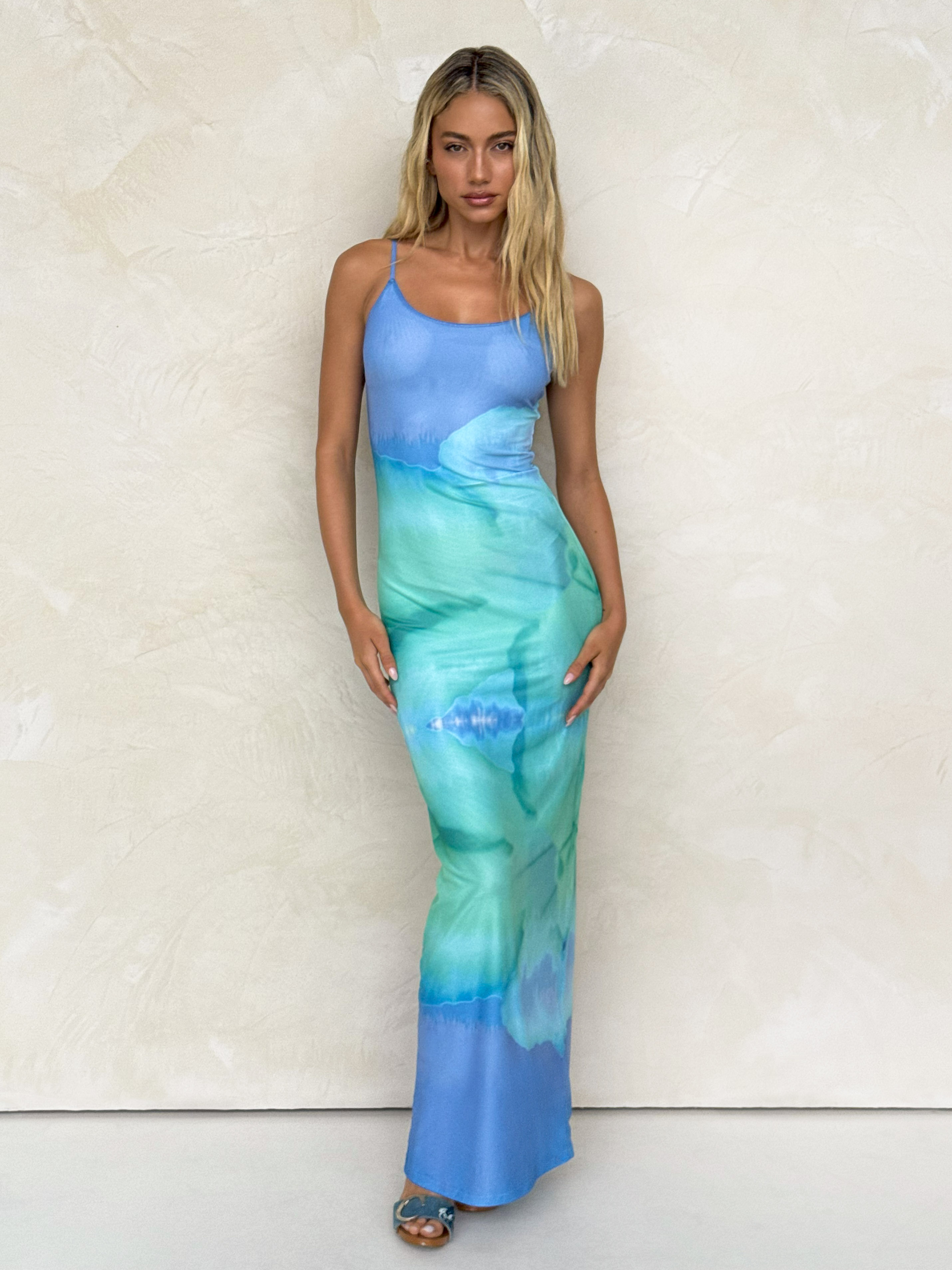 A woman with long blonde hair wears the TIGERMIST ARDON MAXI DRESS in Blue Hibiscus Macro Floral, a form-fitting, sleeveless blue mesh maxi dress, posing against a light textured wall.