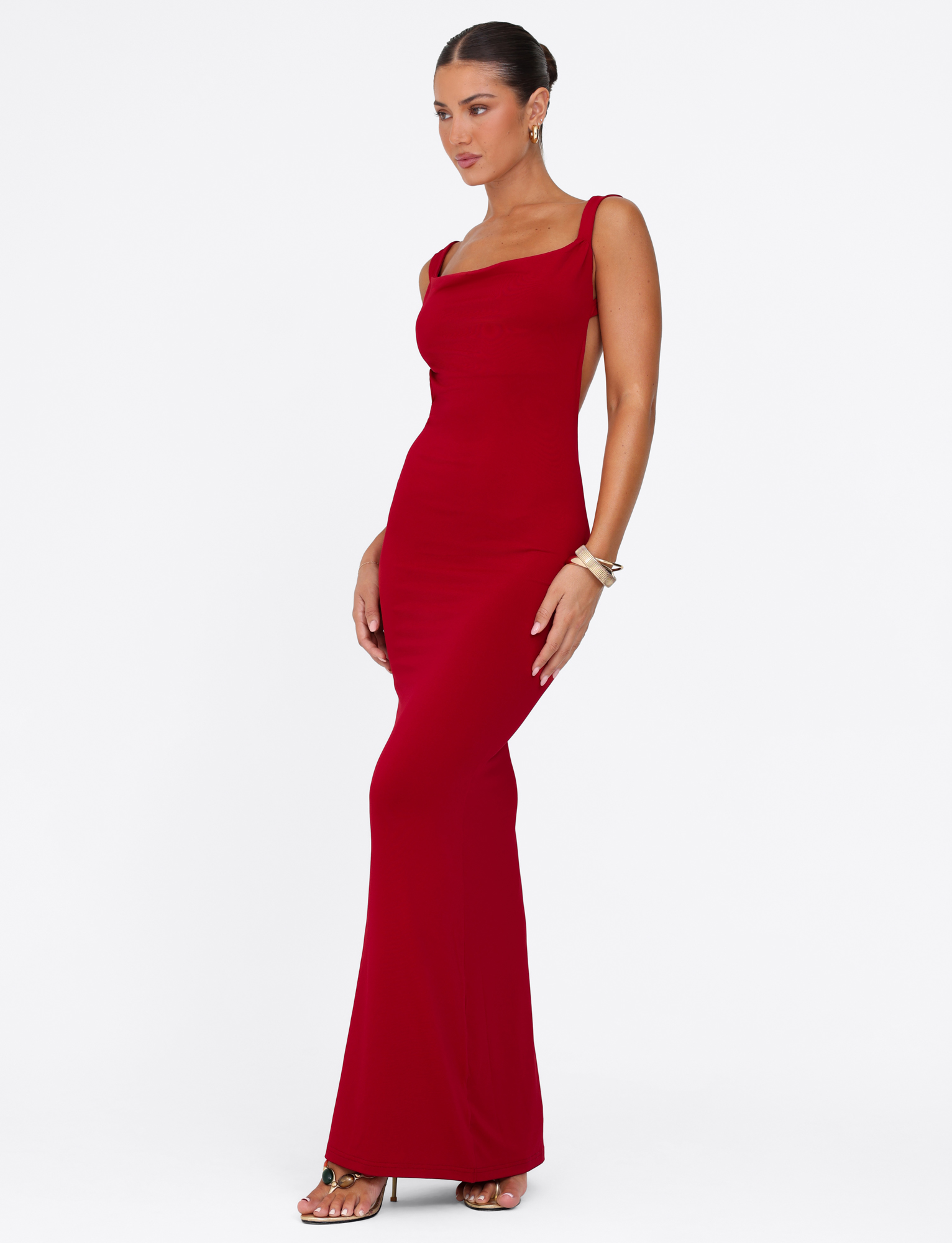 A woman with dark hair in a sleek bun wears the TIGERMIST MORANA MAXI DRESS - RED, a sleeveless, floor-length, bodycon dress, paired with gold sandals and bracelets, standing against a plain white background.
