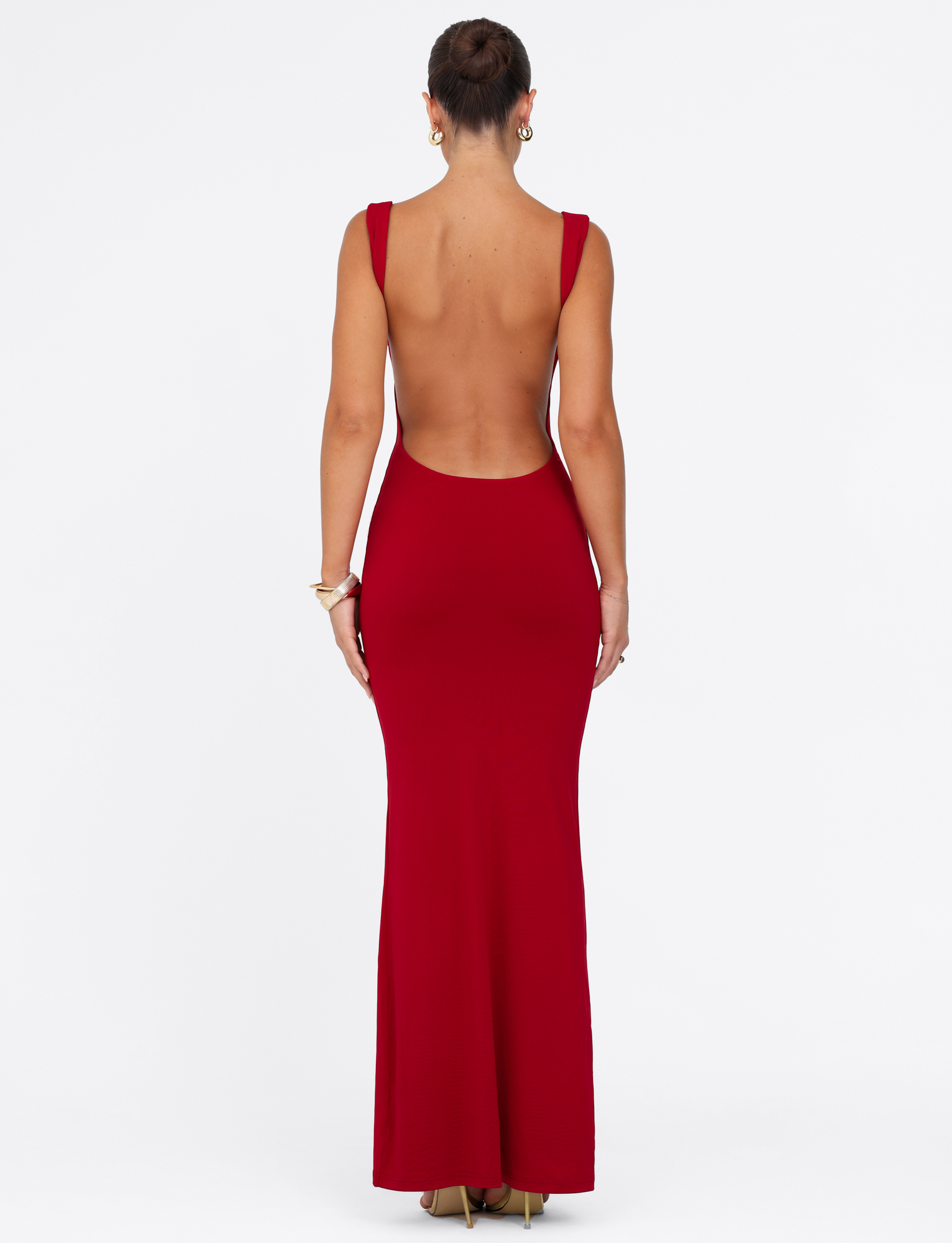 A woman in a sleeveless, backless TIGERMIST MORANA MAXI DRESS - RED, styled with gold heels, bracelets, and hoop earrings, her hair in a bun, poses against a plain white background.