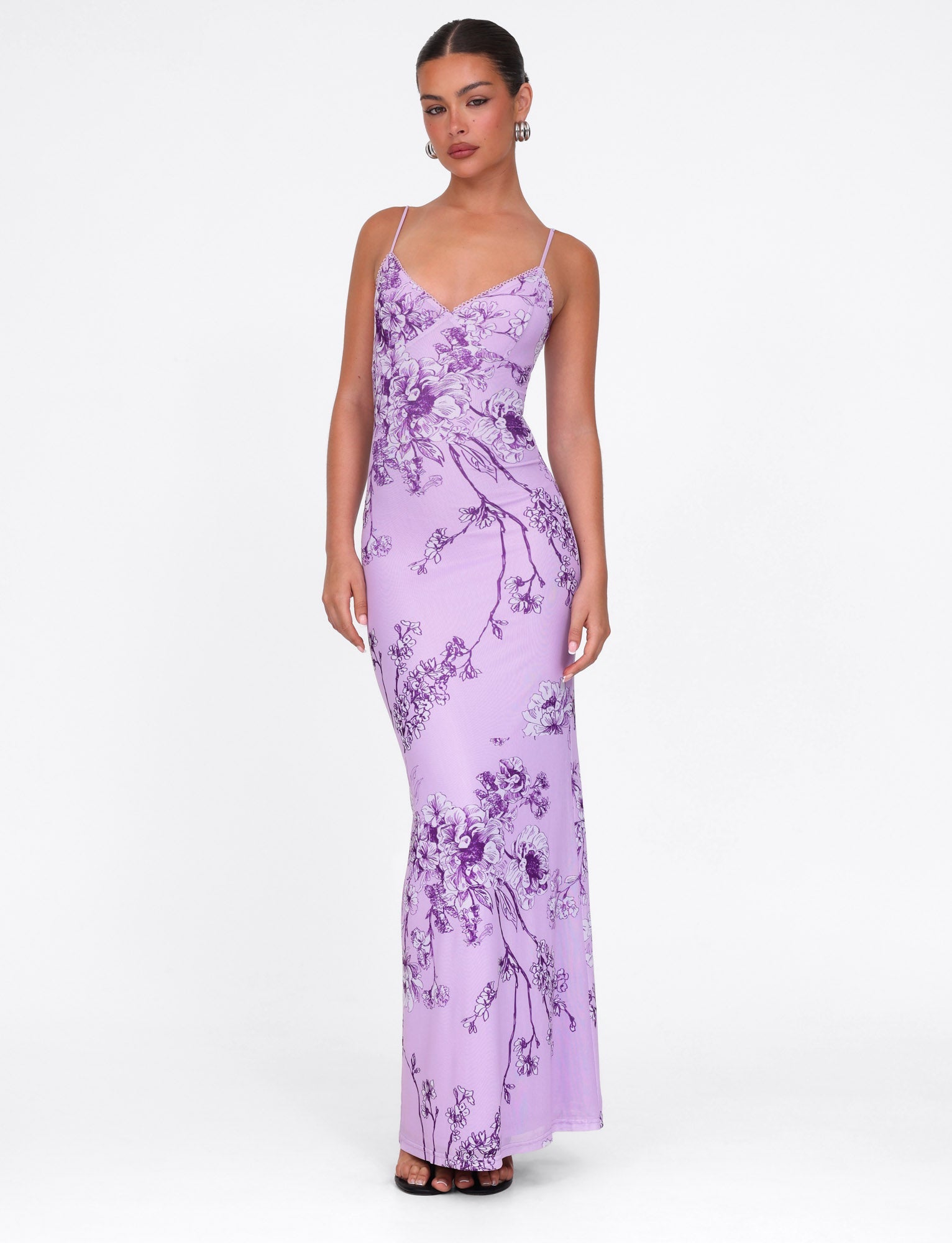 A woman confidently wears the TIGERMIST JULIANNA DRESS in purple ceramic floral, a maxi gown with thin straps, paired elegantly with tied-back hair, hoop earrings, and black heels against a plain white backdrop.