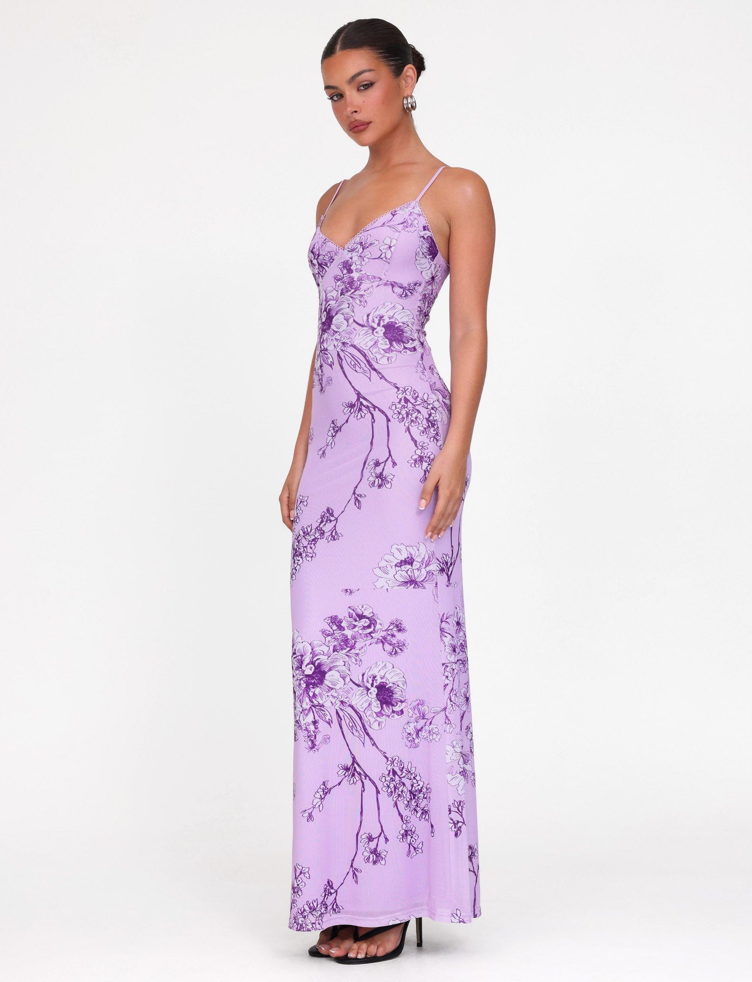A person in a TIGERMIST JULIANNA DRESS - PURPLE : FLORAL : PURPLE CERAMIC FLORAL, maxi-length lilac design, stands against a plain background. Their hair is styled in an updo with elegant earrings and high heels.