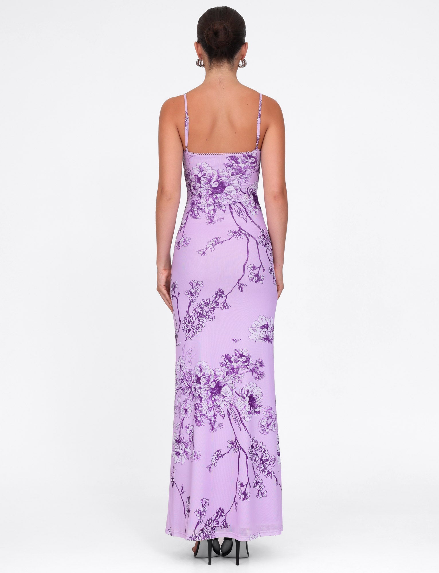 A person is seen from the back wearing a TIGERMIST JULIANNA DRESS, highlighting its maxi length and fitted design. The elegant purple dress features a ceramic floral print and thin straps, standing out against a plain white background.