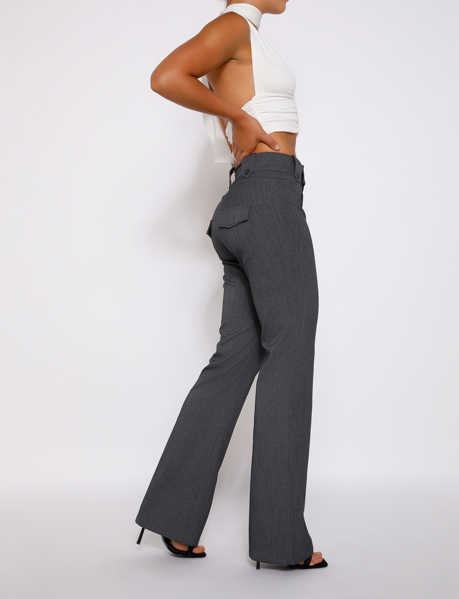 A woman poses in profile against a white background, wearing a sleeveless white crop top with side cutouts, TIGERMIST KITTIE HIGH RISE PANT - GREY with flared legs, and black heeled sandals.