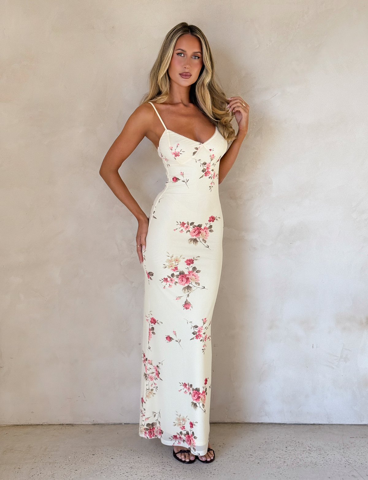 A woman stands against a light wall in the TIGERMIST JULIANNA DRESS in yellow macro pop floral, featuring adjustable spaghetti straps, complementing her long blonde hair and black sandals as she gently touches her hair.