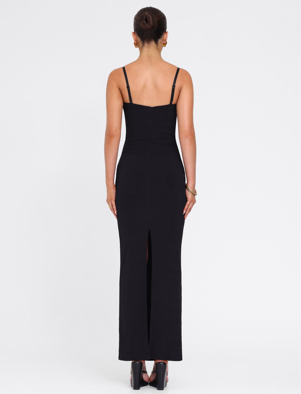 A woman with her hair in a bun faces away, wearing the TIGERMIST ALLURA MAXI DRESS in black—a fitted, sleeveless bodycon style with thin straps and a back slit—paired with high heels against a plain white background.