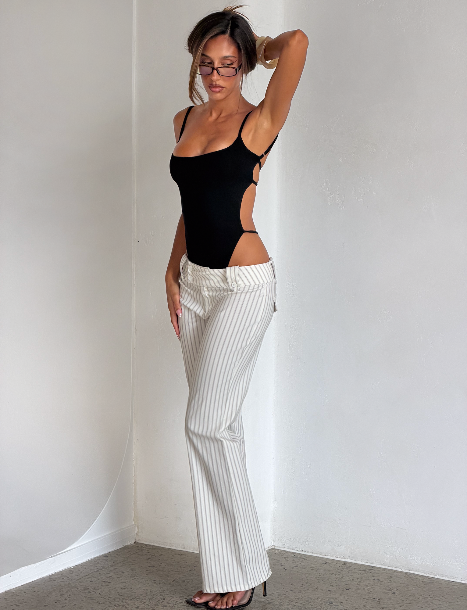 A woman in a black cut-out bodysuit, TIGERMIST KITTIE PANT - IVORY STRIPE, and black heels stands against a white wall, holding her hair up with one hand and wearing rectangular glasses.
