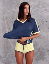 A woman stands against a textured gray wall wearing the TIGERMIST JAYNA TEE in dusty blue and high-waisted yellow drawstring shorts, lifting part of her shirt slightly.