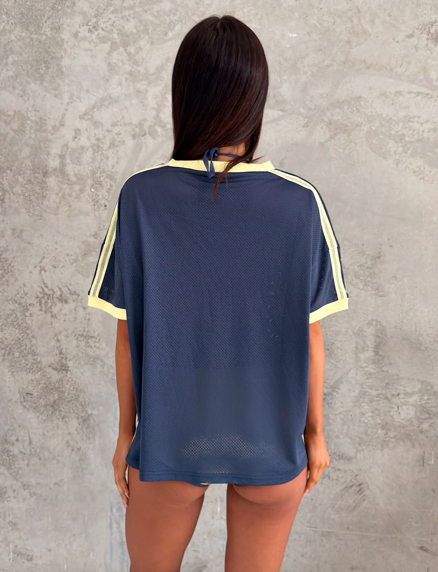 A person with long brown hair stands before a textured gray wall, wearing the TIGERMIST JAYNA TEE in DUSTY BLUE—a loose, slightly sheer sport-style t-shirt with yellow trim and short sleeves.