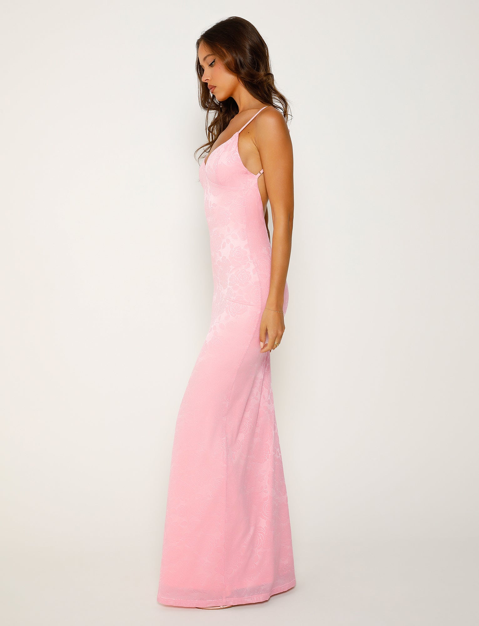 A woman with long brown hair stands in profile against a plain white background, wearing the form-fitting, floor-length ERICKA DRESS in pink by TIGERMIST, featuring thin straps.