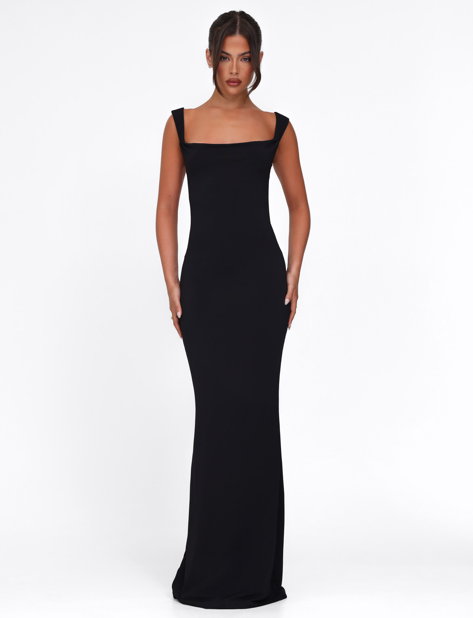 A woman faces forward in the TIGERMIST ZITA DRESS - BLACK, a bodycon maxi with wide shoulder straps. Her hair is in a neat updo as she poses against a plain white background.
