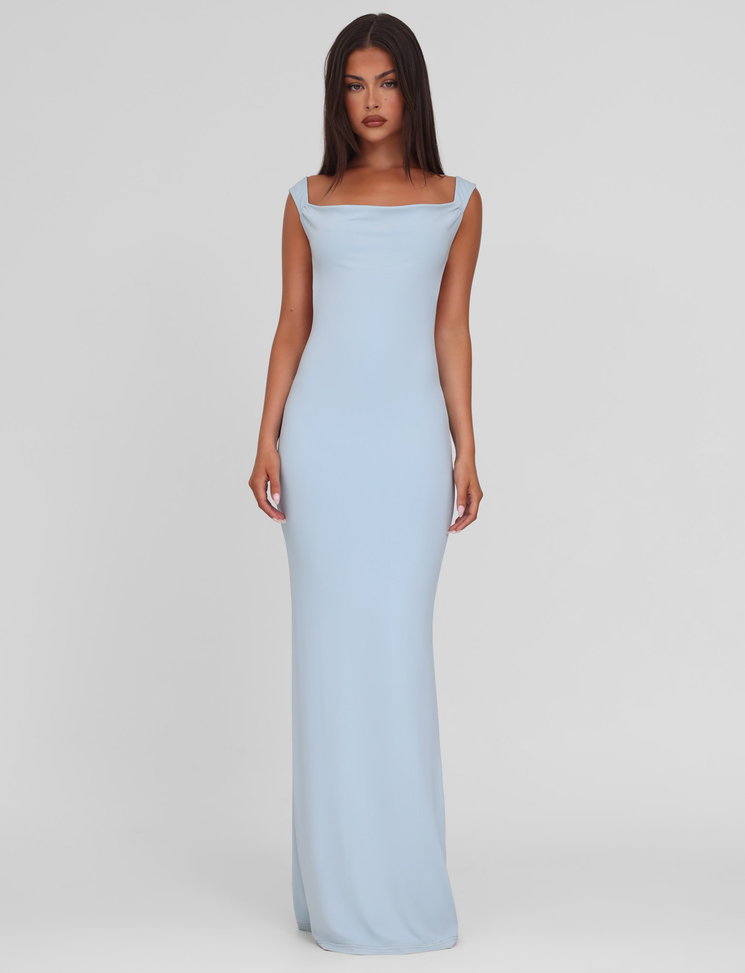 A woman wears the TIGERMIST ZITA DRESS - BLUE in baby blue, a sleeveless, floor-length bodycon gown with a square neckline, standing against a plain white background.