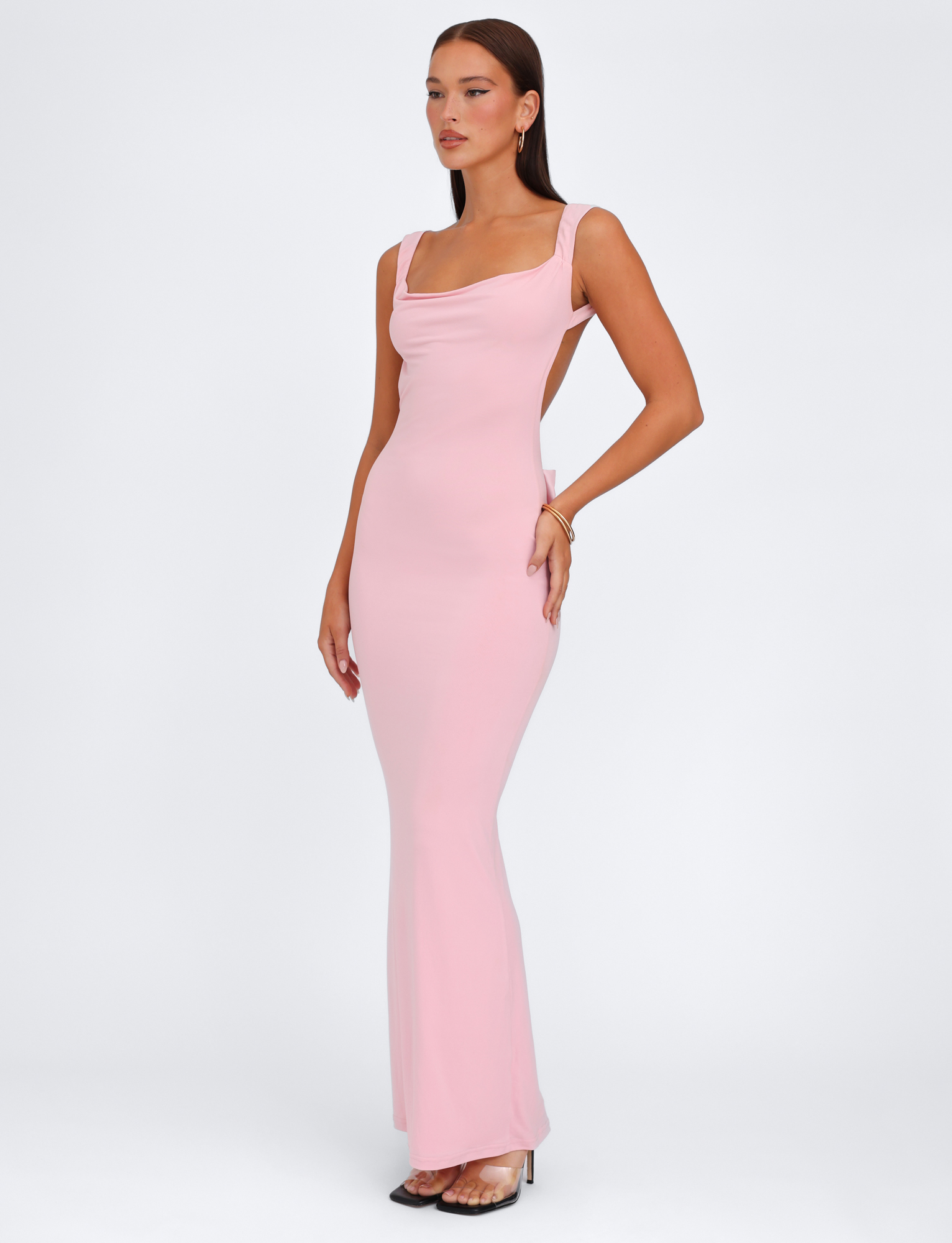 A woman models the TIGERMIST ZITA DRESS - PINK : BABY PINK, showcasing its bodycon fit and square neckline. She pairs it with clear heels and gold jewelry, standing against a plain white background.