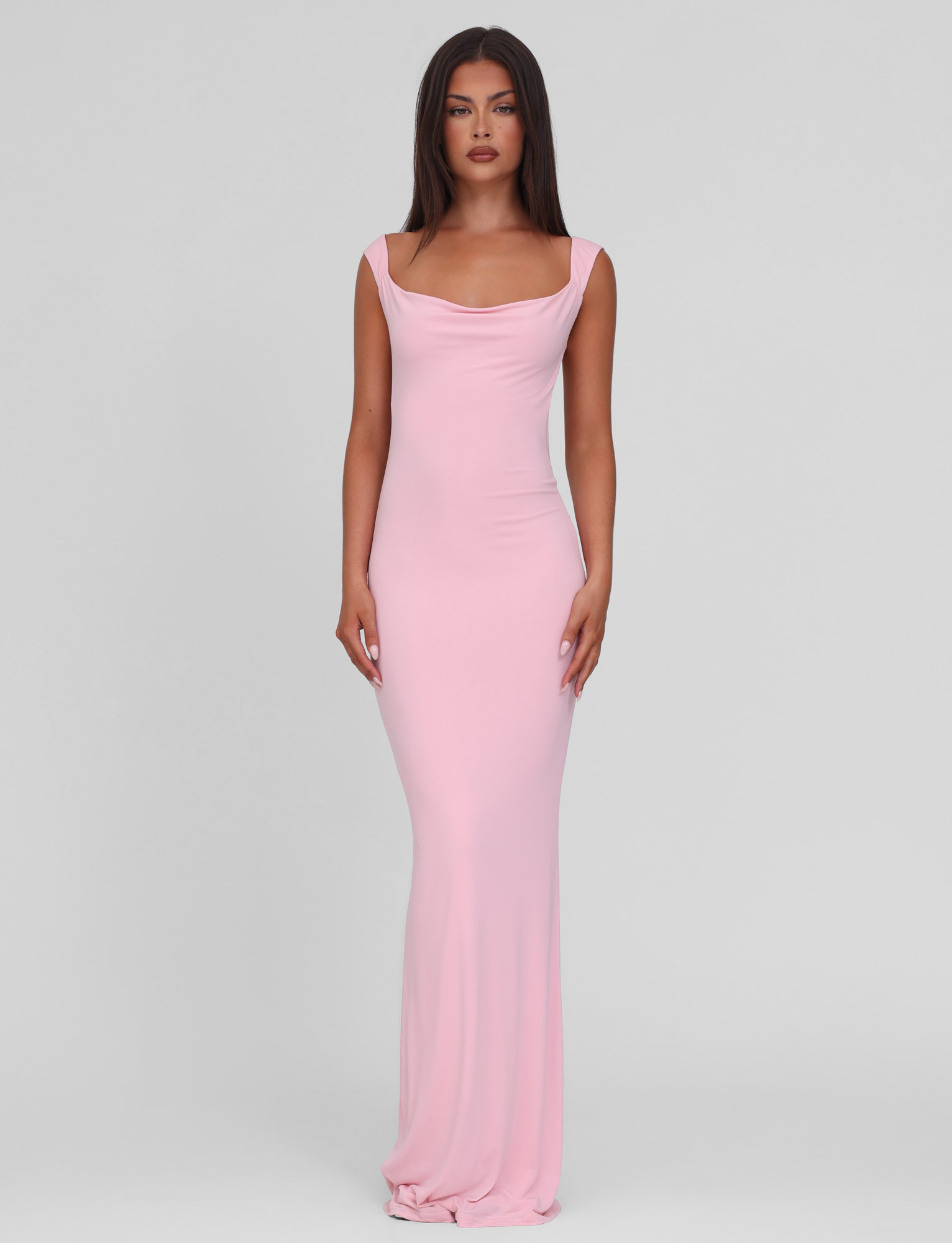 A woman faces forward, wearing the TIGERMIST ZITA DRESS in baby pink—a fitted, sleeveless bodycon maxi dress with a draped neckline—against a plain light gray background.