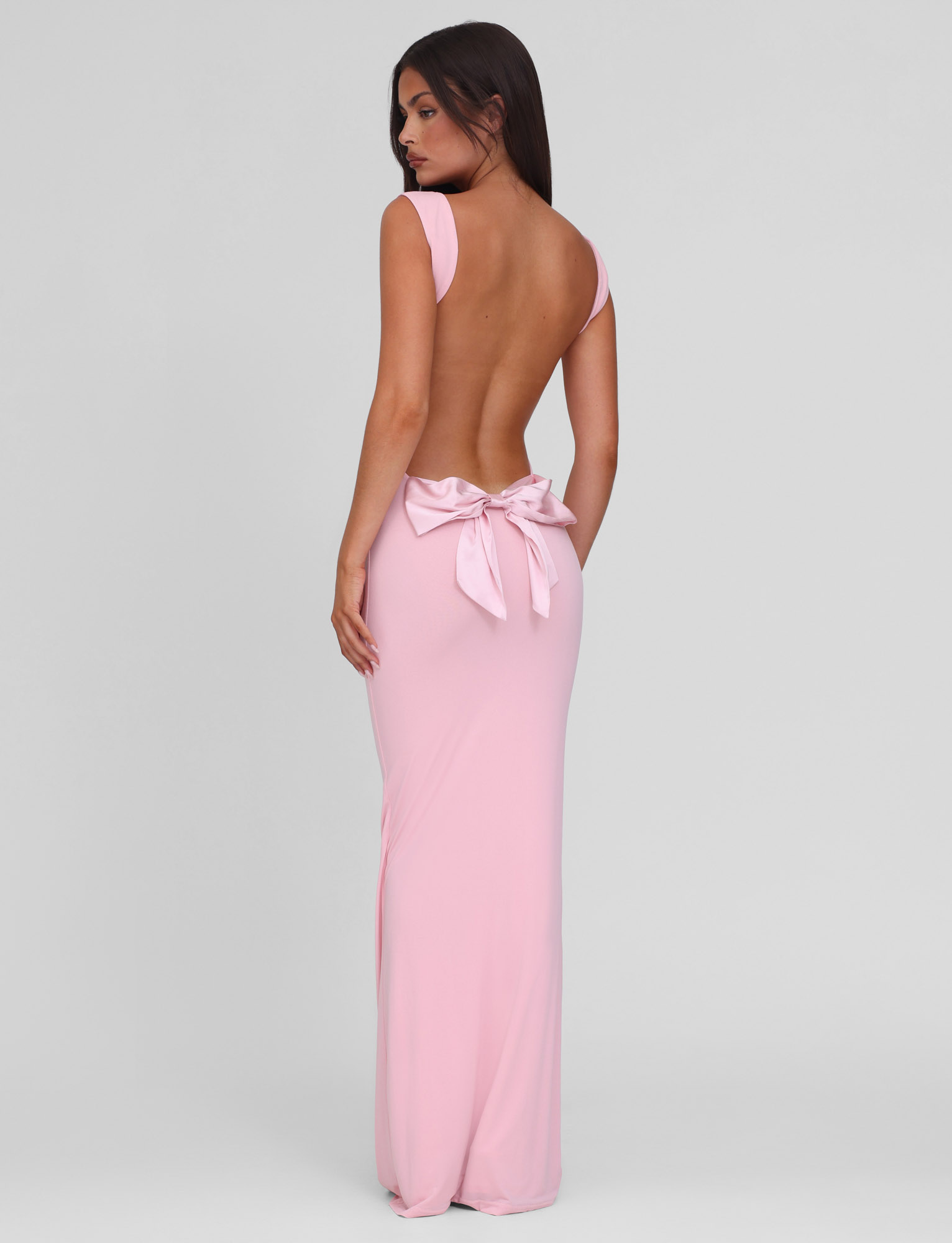 A woman in the TIGERMIST ZITA DRESS - PINK : BABY PINK, a long maxi dress with a large back bow, stands against a light gray background and looks over her shoulder.