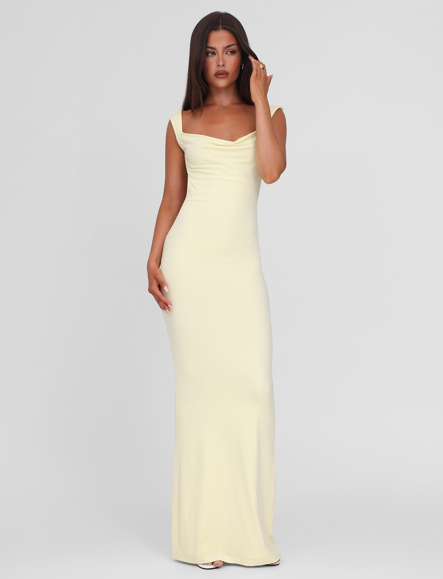 A woman with long dark hair stands against a plain background wearing the TIGERMIST ZITA MAXI DRESS - LEMON, a sleeveless light yellow bodycon gown with a scoop cowl neck. She touches her face and gazes slightly to the side.