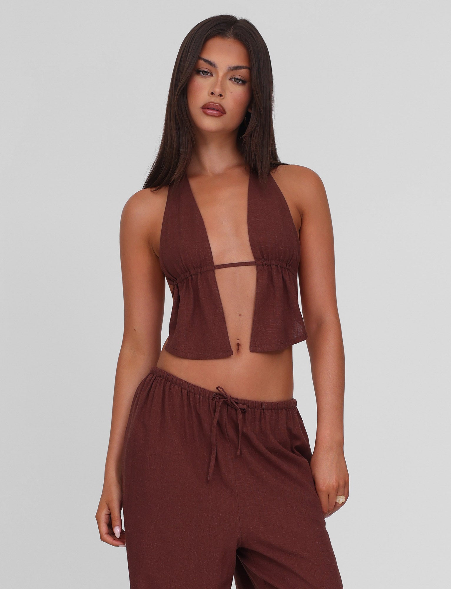 A woman with long dark hair wears the TIGERMIST LAINEY TOP in chocolate, a linen halter top with a deep V-neck, paired with matching drawstring pants, standing against a plain light background.