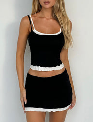 A woman with long blonde hair is wearing the ADAIR TOP - BLACK : BLACK/WHITE from TIGERMIST, paired with a matching black mini skirt with white trim. She is standing against a plain white background.