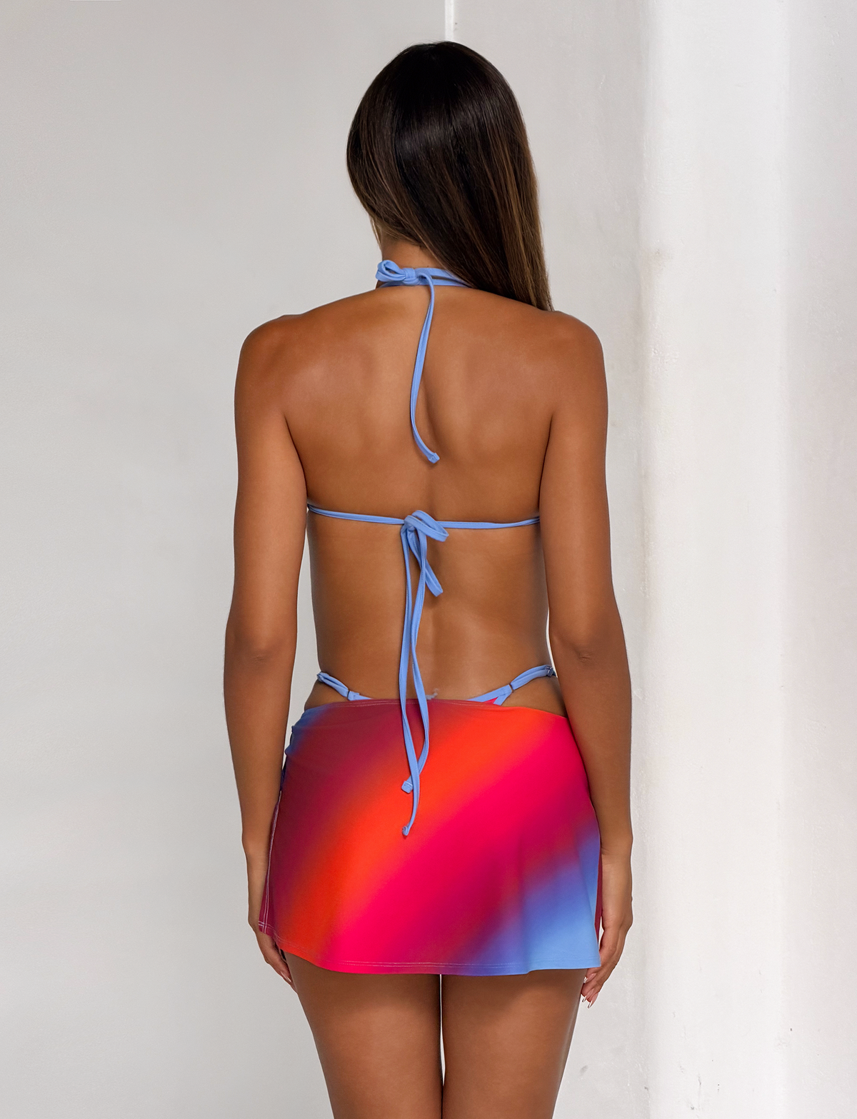 A woman with long brown hair stands indoors by a white wall, wearing the TIGERMIST DIANA BIKINI TOP in CIELO OMBRE with a short, colorful red, pink, and blue ombre stripe skirt—her back faces the camera.
