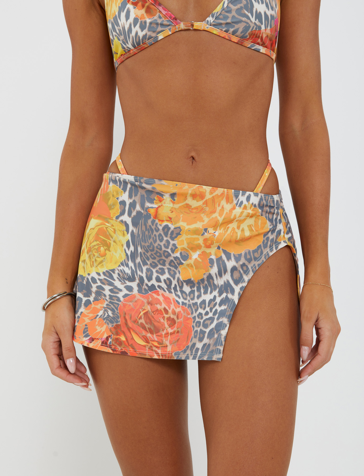 A woman wears the TIGERMIST DIANA TWO-PIECE BIKINI BOTTOM in Orange Leopard, styled with a matching high-slit swim skirt featuring vibrant orange leopard and floral patterns in yellow, orange, and gray.