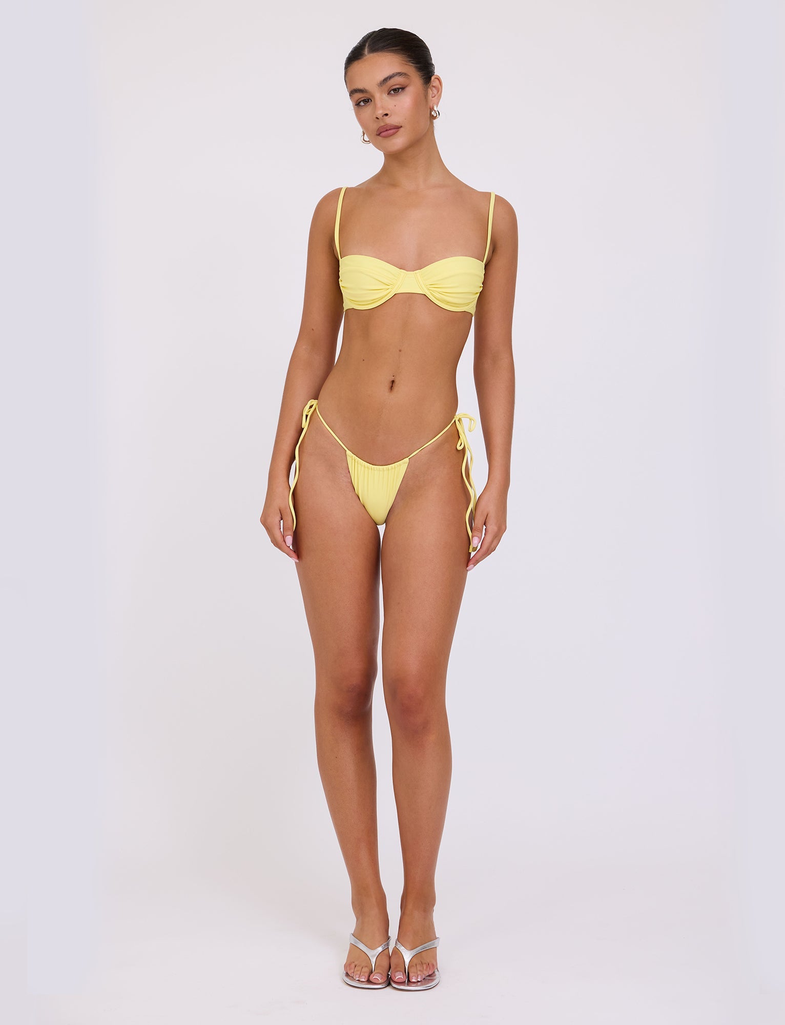 A person stands confidently, facing the camera, wearing a yellow TIGERMIST bikini with thin straps. The top features a ruched design, and they are sporting the KHESSA BIKINI BOTTOM - YELLOW : LEMON tied at the hips. They also wear white sandals and have their hair pulled back. The background is a plain white.