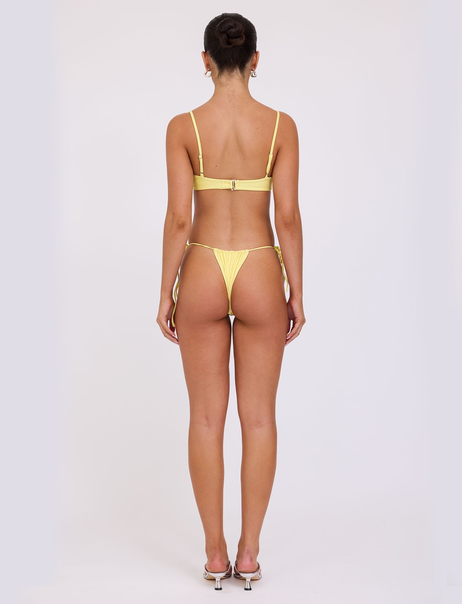 A woman is standing with her back to the camera, wearing a TIGERMIST KHESSA BIKINI BOTTOM - YELLOW : LEMON. Her hair is tied up in a bun, and she is also wearing high-heeled sandals. The image shows her full body from head to feet against a plain white background.