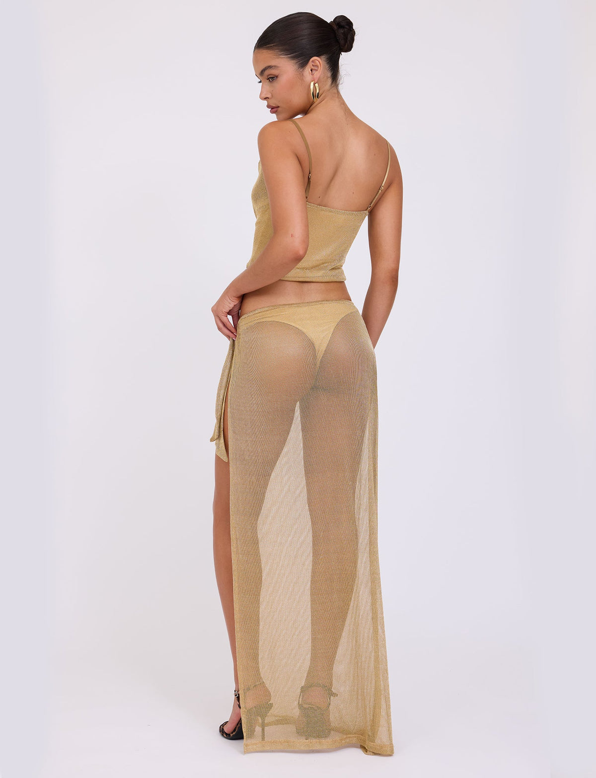 A person with their hair in a bun stands facing away from the camera, wearing TIGERMIST's WYNNE TOP in gold, paired with a matching long skirt that reveals their undergarments. They have complemented this look with black strappy heels and gold hoop earrings.