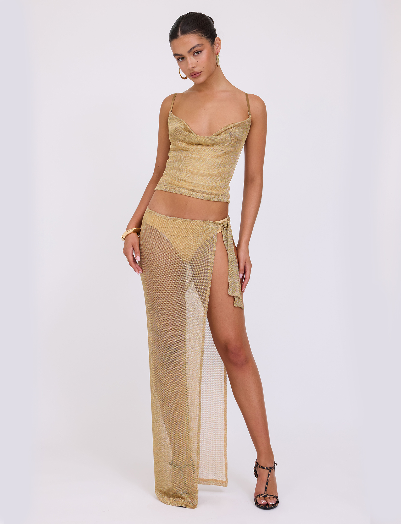 A woman models the WYNNE SKIRT - GOLD by TIGERMIST, a sheer gold two-piece set with a sleeveless top and high-slit skirt. She accessorizes with hoop earrings and black heeled sandals against a plain white background.