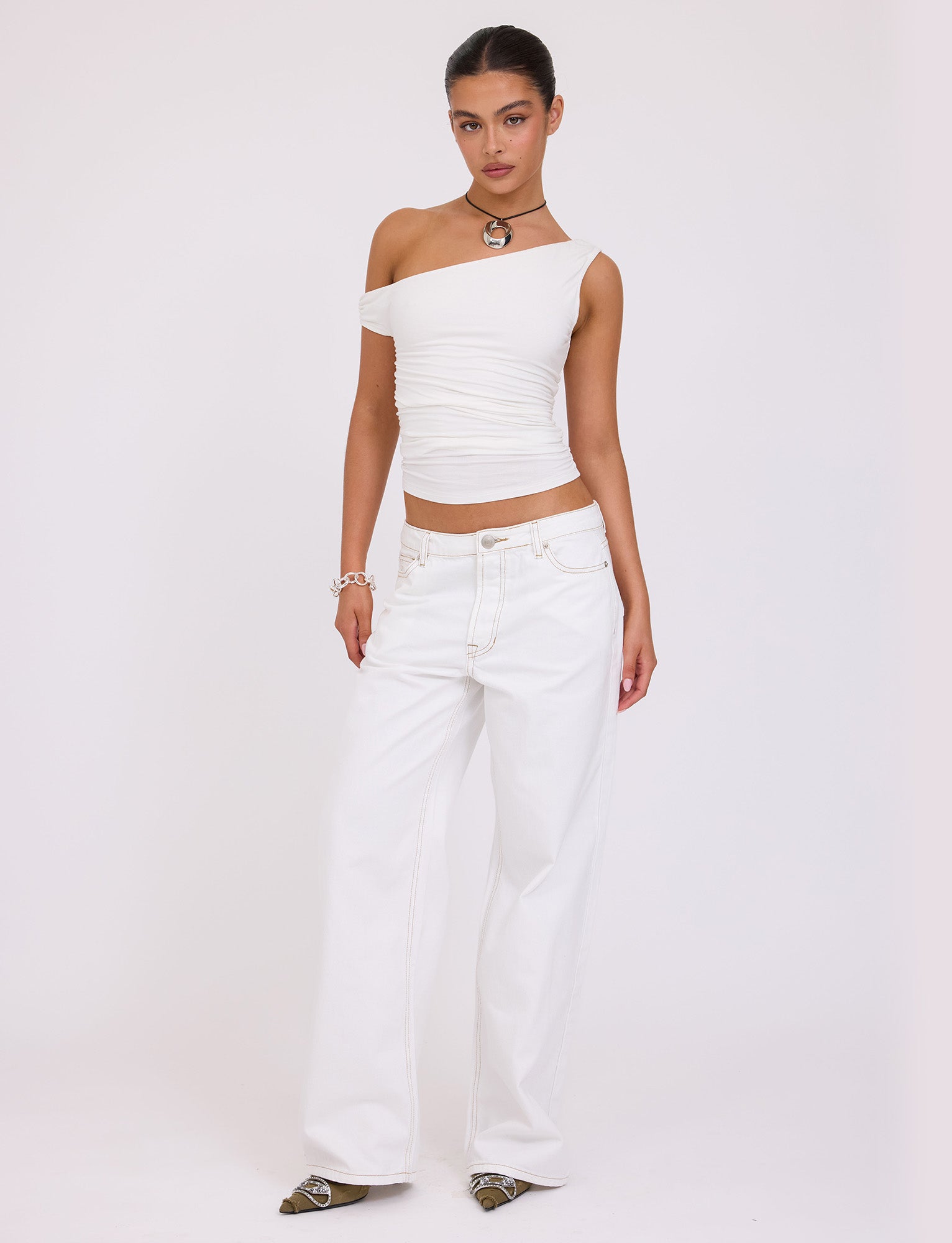 A person stands against a plain white background, wearing the asymmetrical FELIX JEAN - WHITE top and wide-leg jeans from TIGERMIST. They accessorize with a necklace, bracelet, and pointed-toe shoes. Their hair is pulled back as they pose with one hand on their hip.