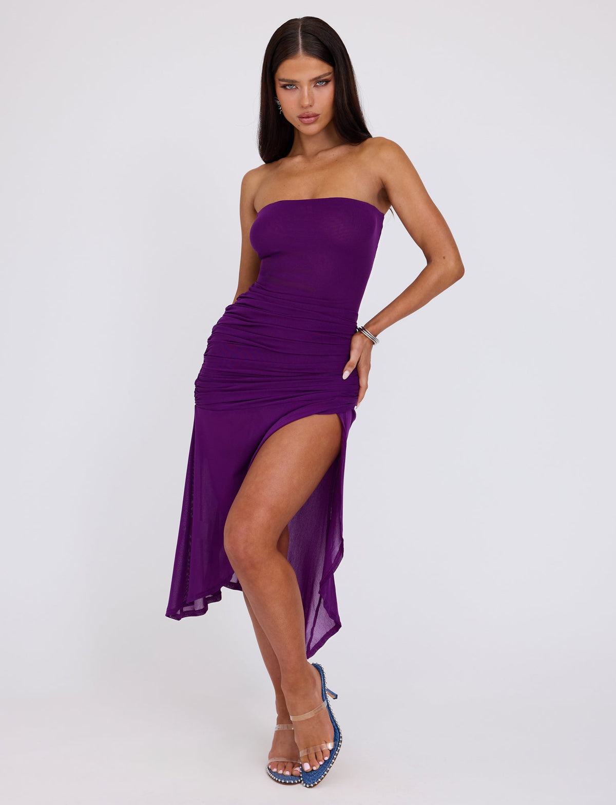 A woman with long dark hair poses against a white background, wearing the COVE DRESS - PURPLE by TIGERMIST, featuring a strapless design with ruching and an asymmetrical hemline. The dress also includes a high slit on the left side. She completes the outfit with clear, studded high-heeled sandals.