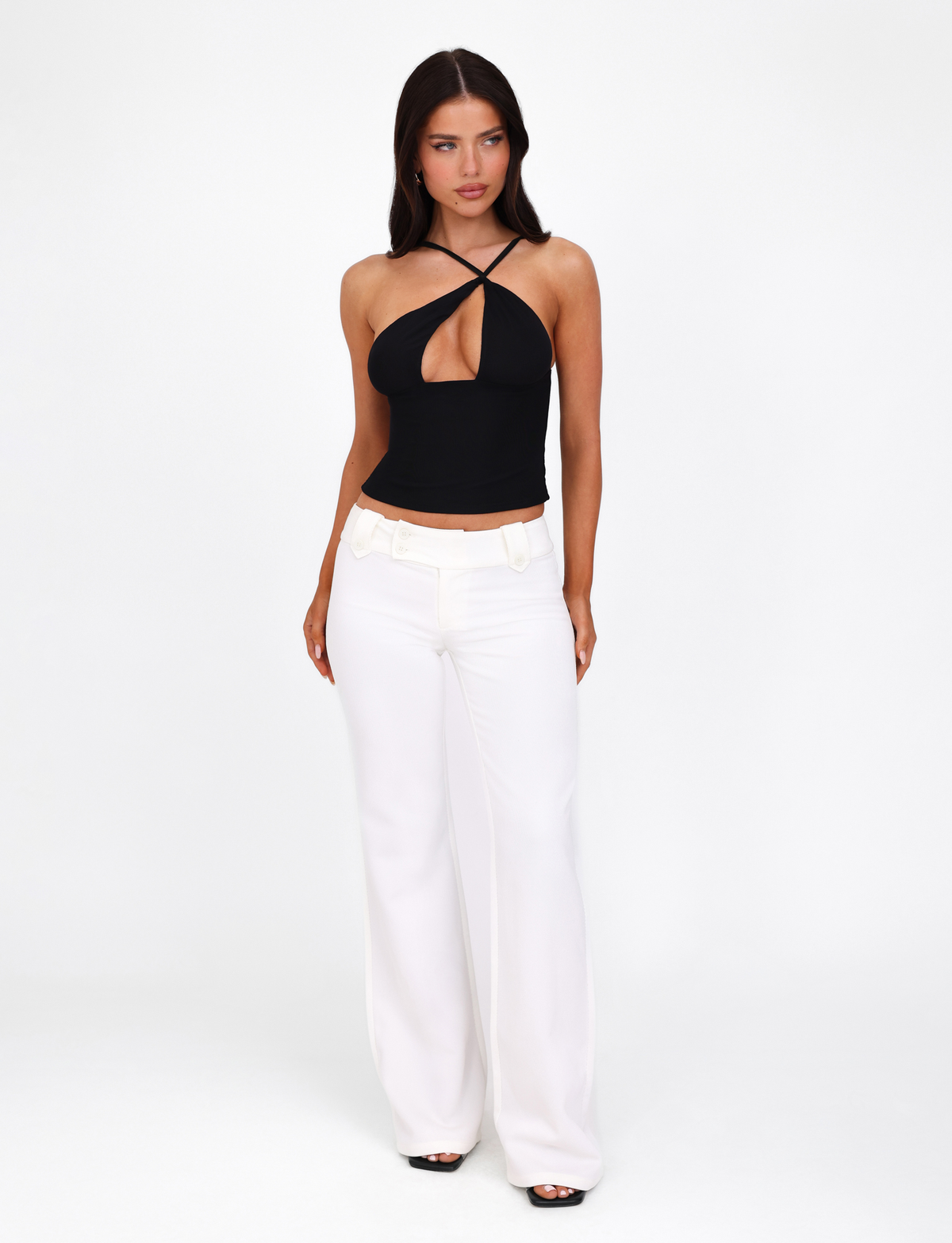 A woman with long dark hair models the TIGERMIST ACADIA TOP in black, featuring an asymmetric neckline, paired with high-waisted white wide-leg pants. She stands against a plain white background, gazing slightly to the side with her hands at her sides.