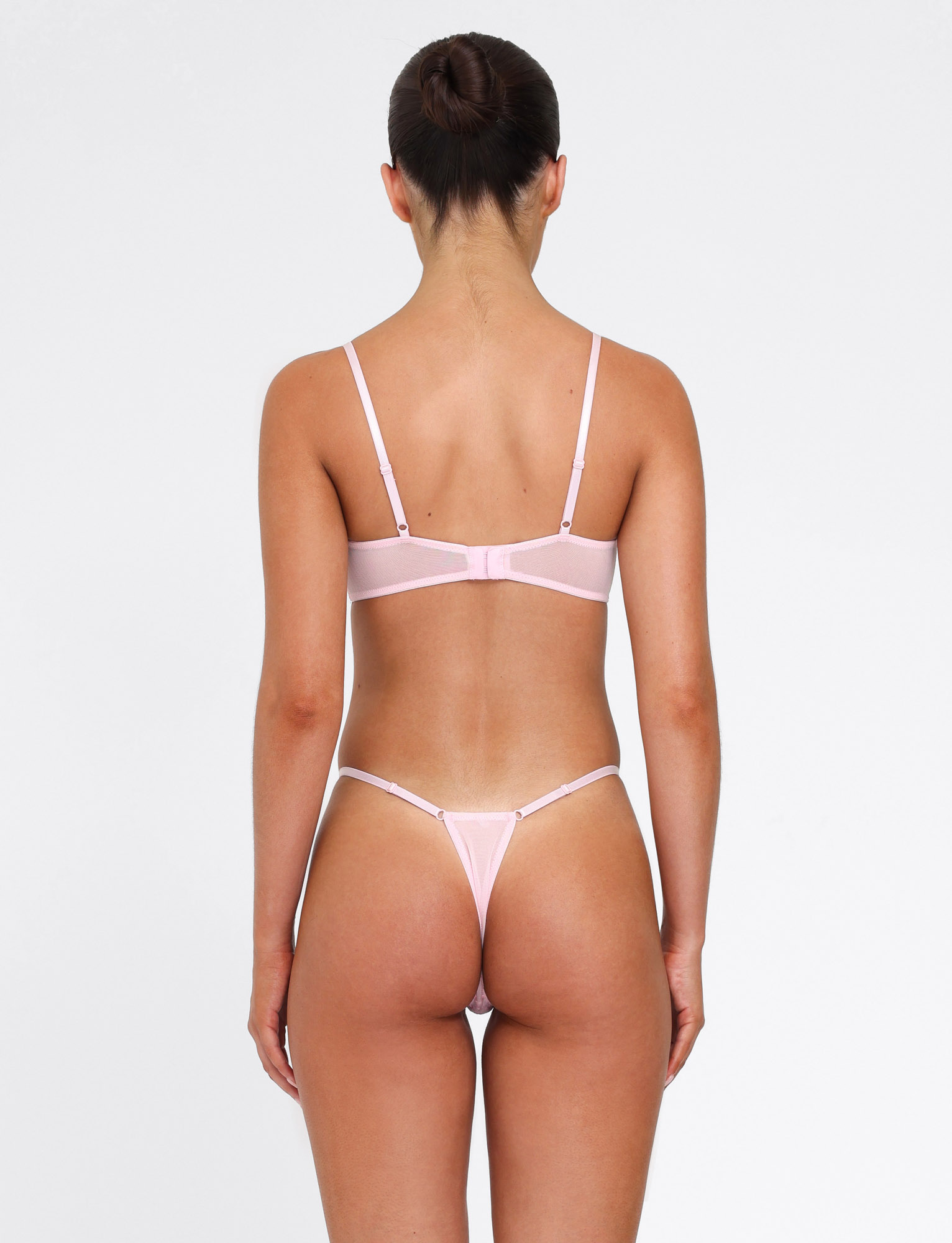 A woman with dark hair in a bun stands with her back to the camera, wearing the TIGERMIST TALIYAH SET - PINK : BABY PINK bra and g-string against a plain white background.